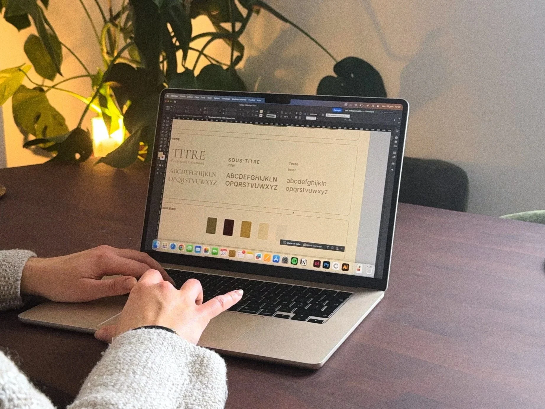 Une personne travaillant sur un ordinateur portable avec un logiciel de conception graphique sur un bureau en bois, arrière-plan un mur clair et une plante verte.