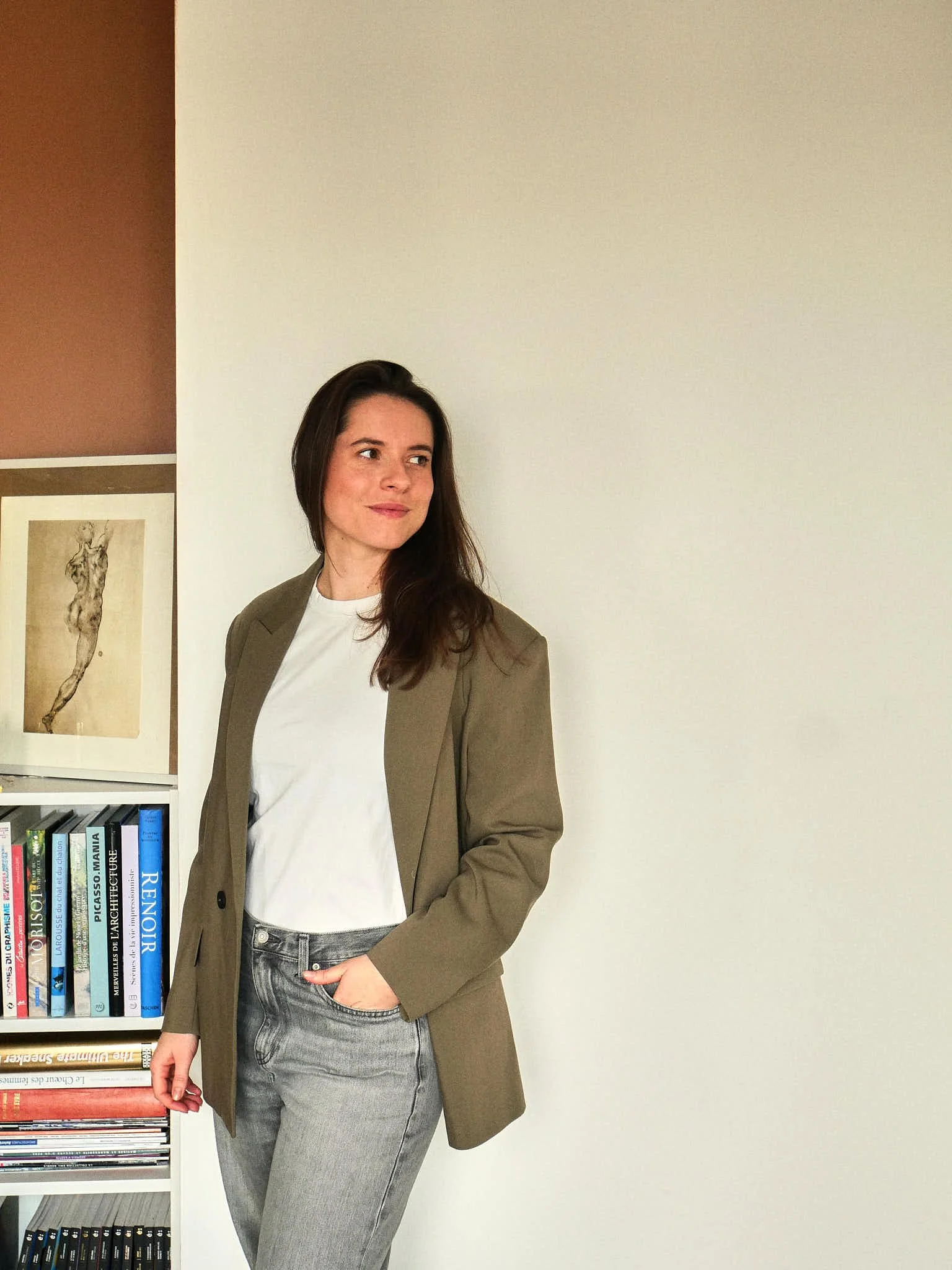 Femme debout à côté d'une étagère remplie de livres, portant une veste beige, un t-shirt blanc et un jean gris, apparemment posant ou regardant vers la gauche