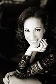 Black-and-white portrait of a smiling woman with dark, curly hair, wearing a lace top, resting her chin on her hand.