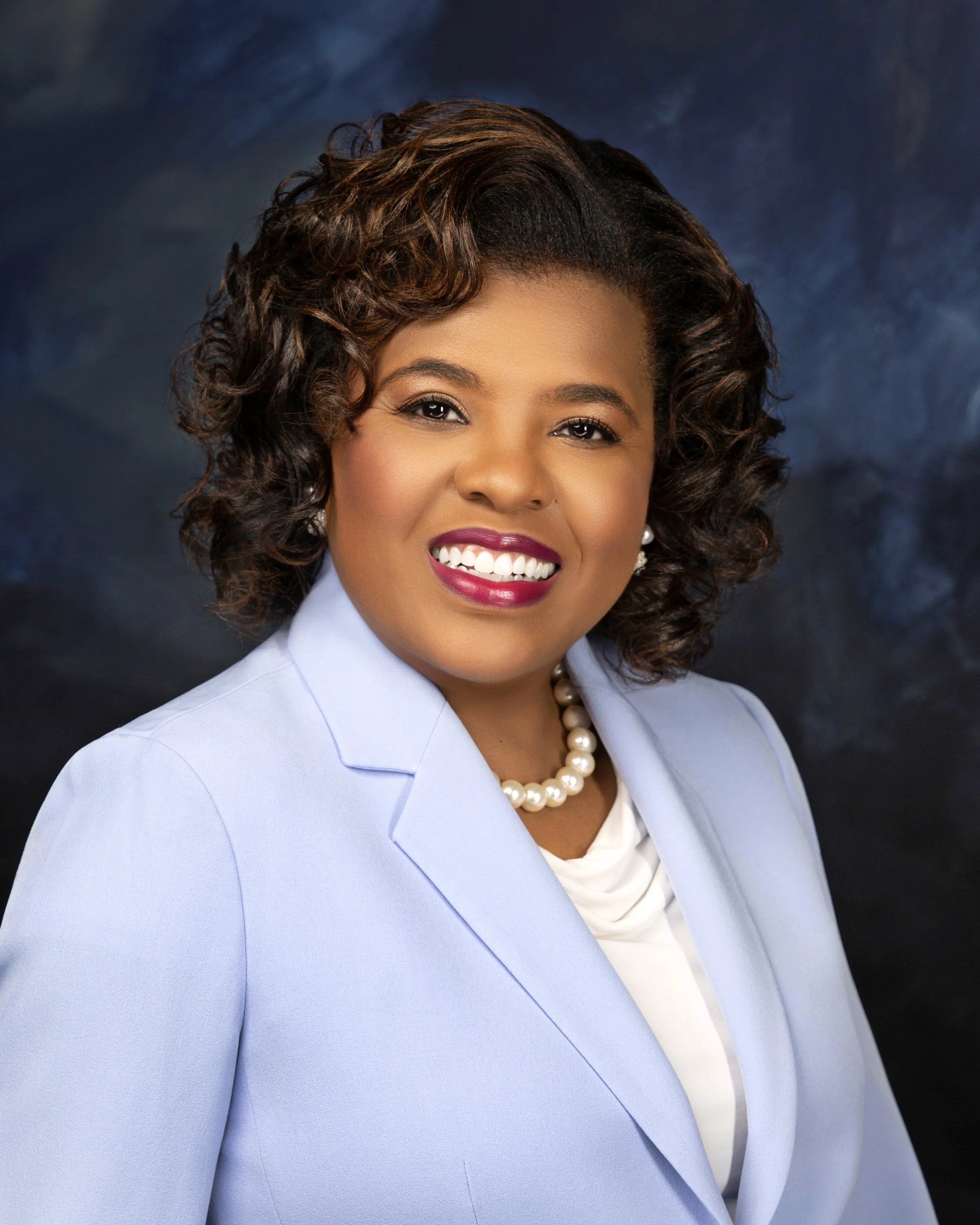 A close-up portrait of a woman with curly brown hair, wearing a light blue blazer, white blouse, pearl necklace, and makeup with dark pink lipstick, smiling against a dark, cloudy background.