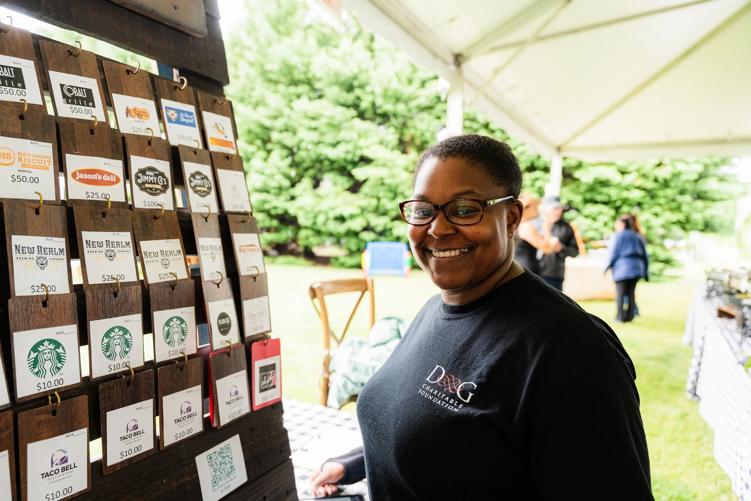 Smiling woman wearing glasses and a charity t-shirt at an outdoor event, standing next to a display of cards and price tags for various businesses.