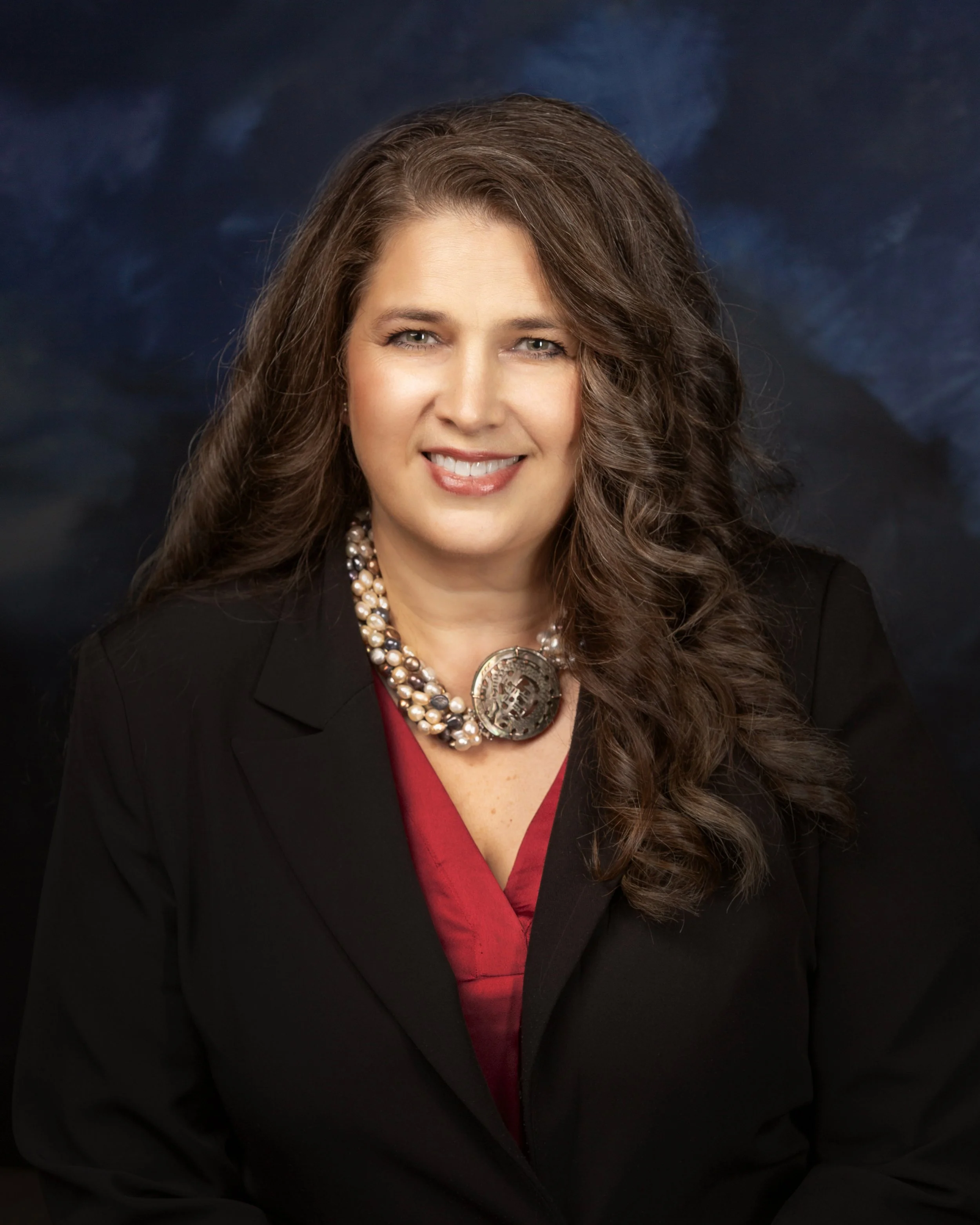 A woman with long, curly brown hair, wearing a black blazer with a red shirt underneath, featuring a pearl necklace and a large decorative pendant. She has light skin, a bright smile, and is posing against a dark, textured background.