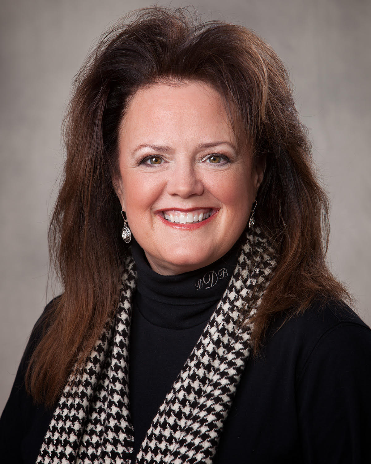 A woman with shoulder-length reddish-brown hair, smiling, wearing silver earrings, a black turtleneck with embroidered initials, and a black and white houndstooth scarf against a neutral background.