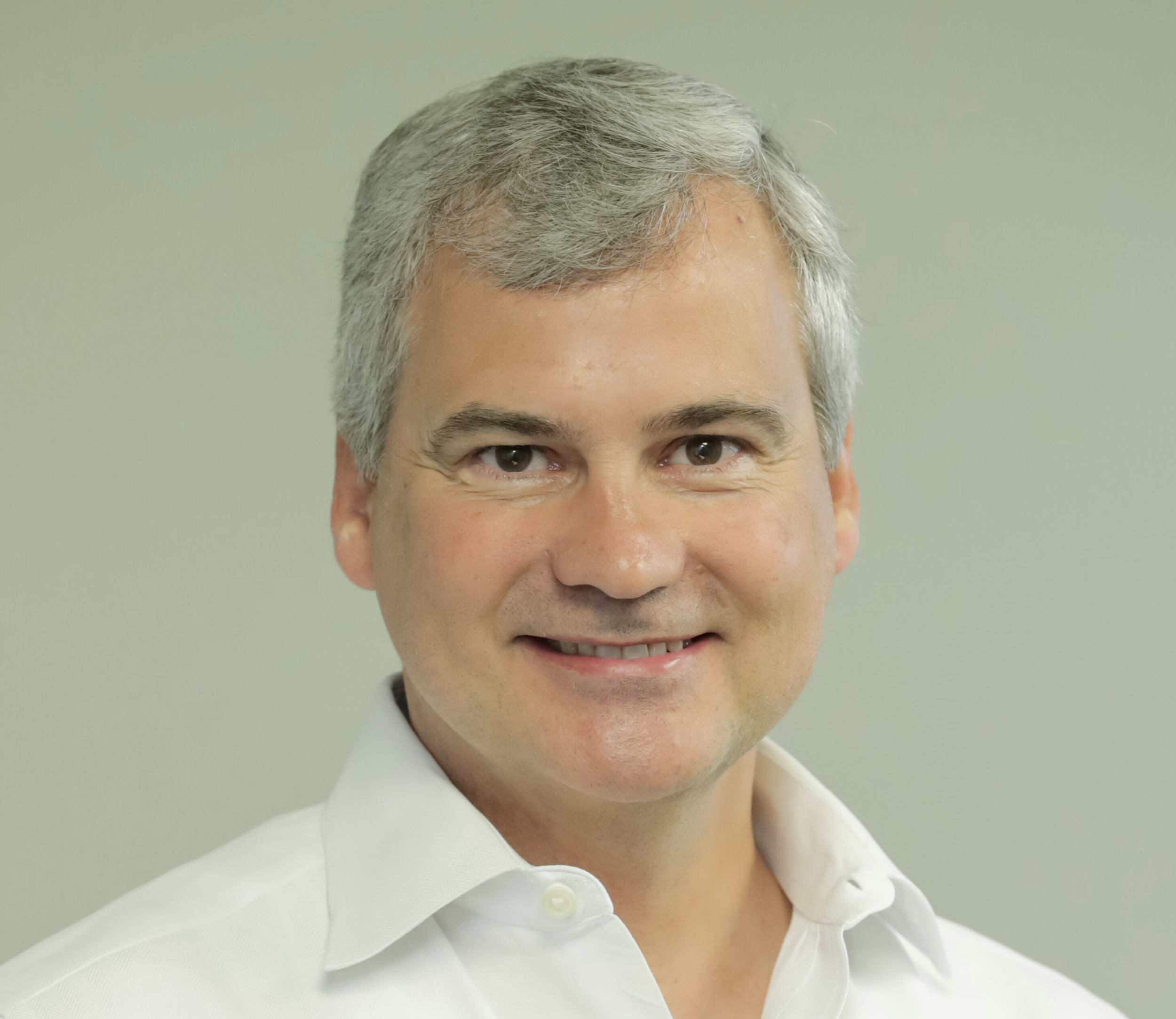 Close-up of a middle-aged man's face, smiling, with gray hair and wearing a white collared shirt, against a light green background.