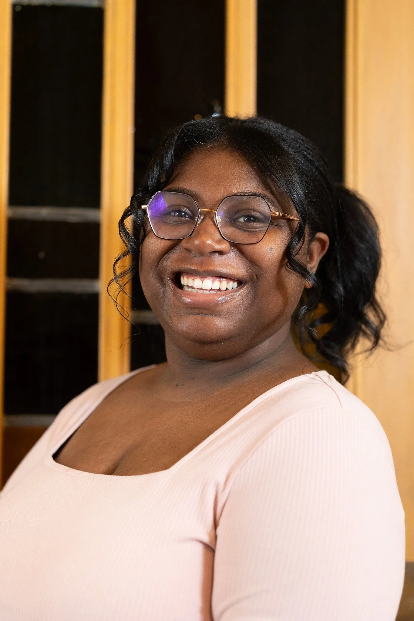 A woman smiling, wearing glasses and a light pink top, with a wooden cabinet in the background.