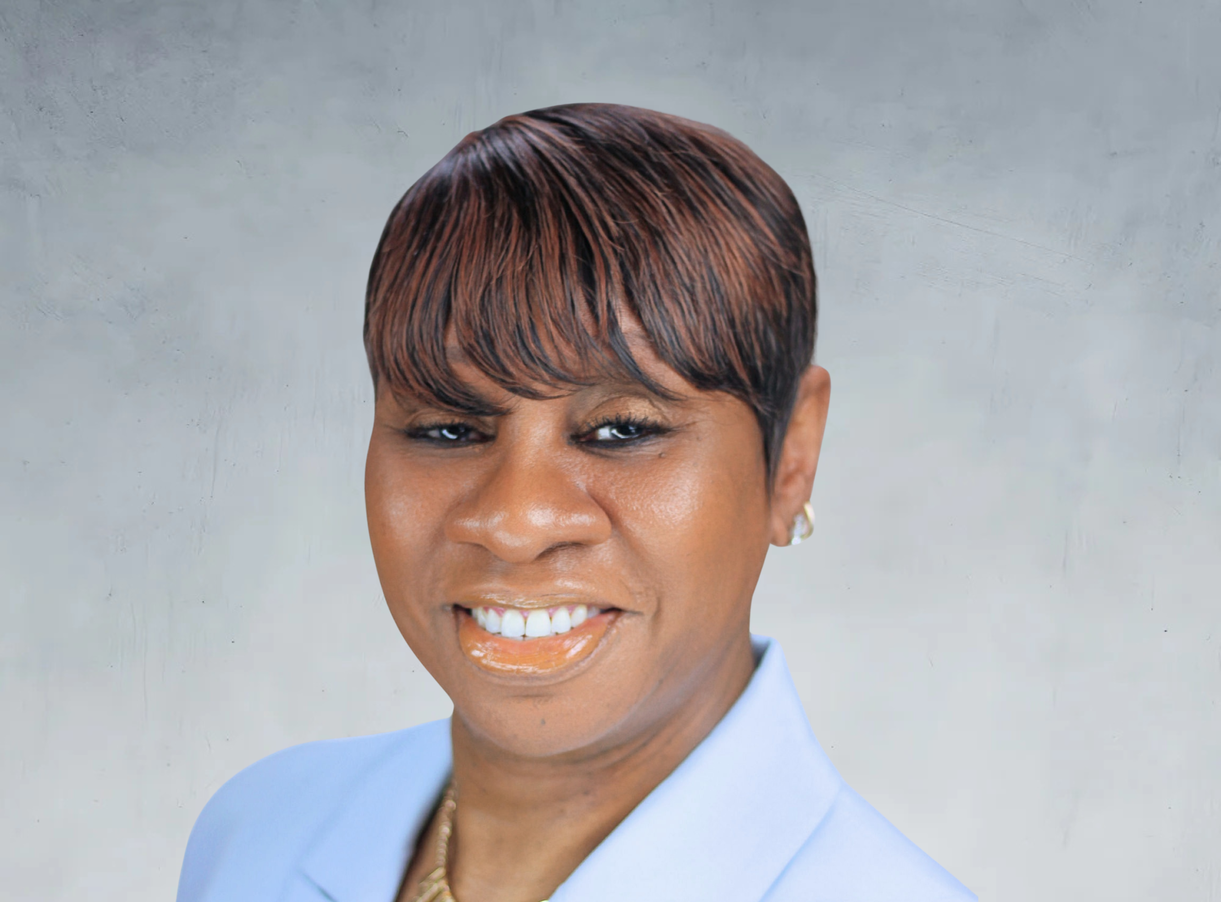 A woman with a short, layered hairstyle with auburn highlights. She is smiling, wearing light makeup, a light blue blazer, and gold jewelry, standing against a plain light gray background.