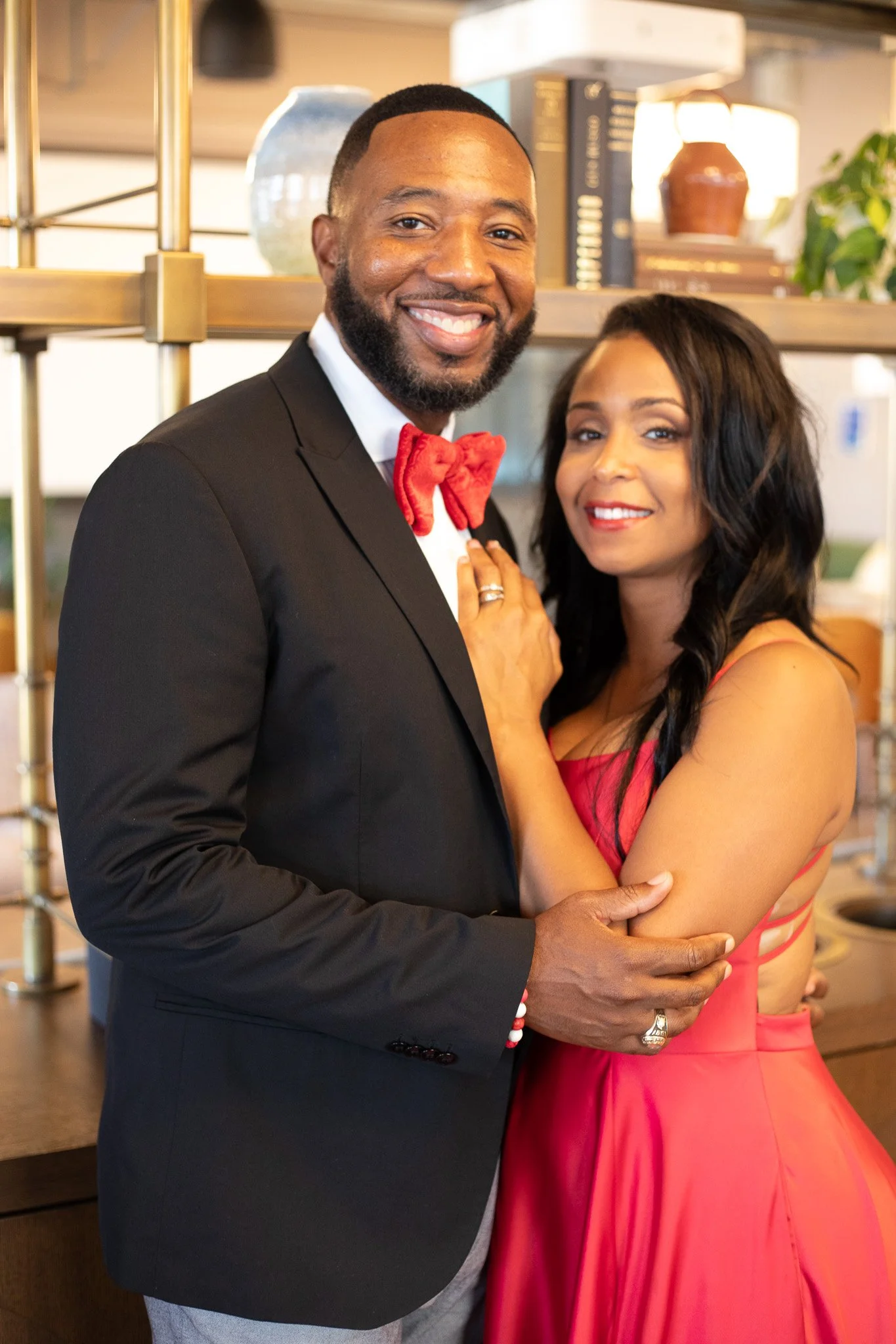 A smiling couple dressed in formal attire stands indoors, with the man wearing a black tuxedo and red bow tie, and the woman in a red dress. They are embracing each other.