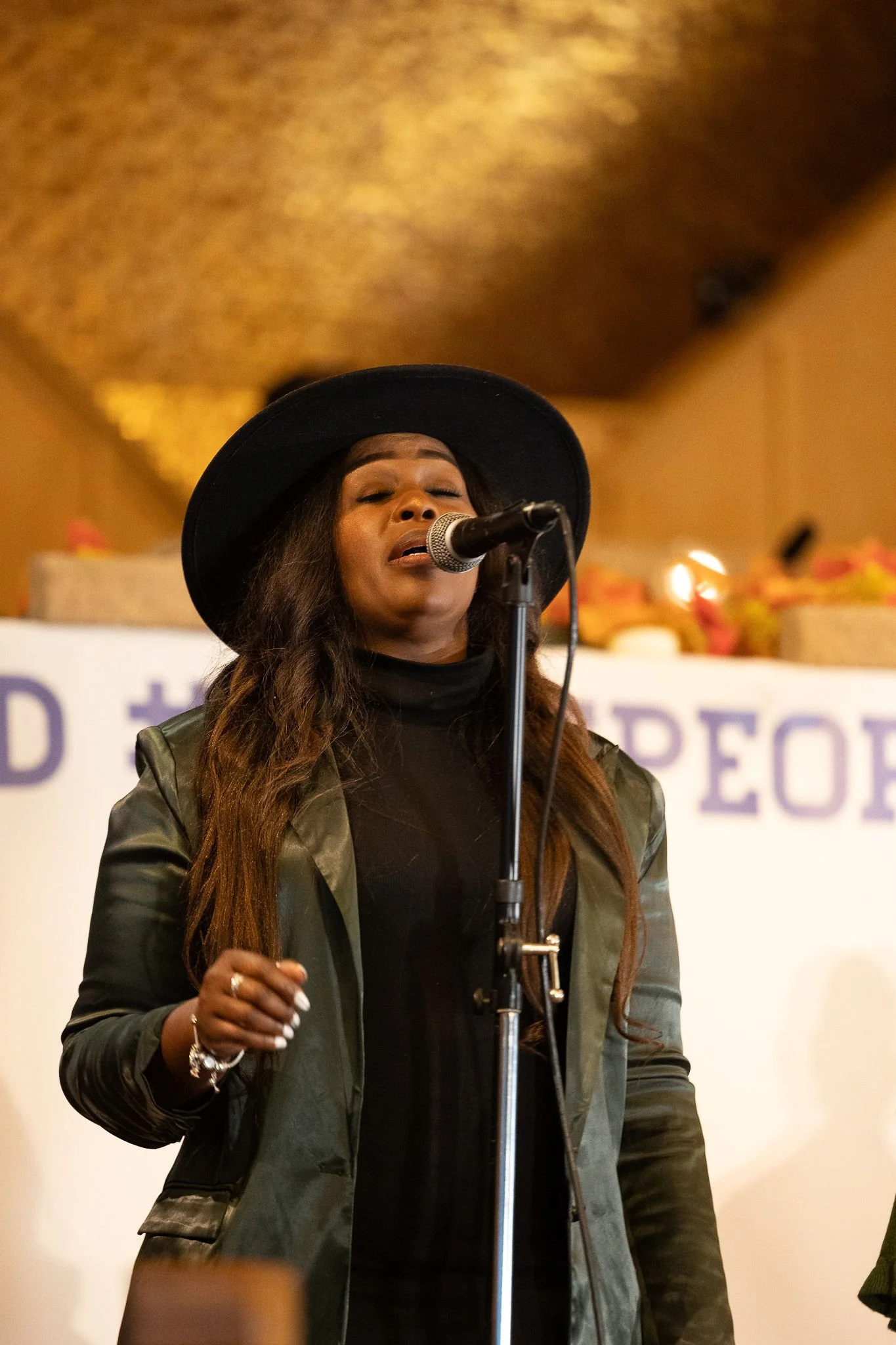 A woman singing into a microphone on stage, wearing a black hat and a green jacket, with a blurred background that includes a banner and some decorations.