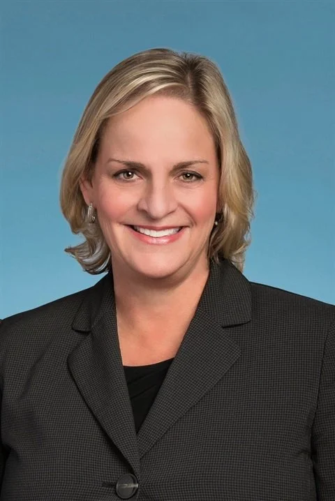 A professional woman with blonde hair in a gray business suit smiling against a blue background.