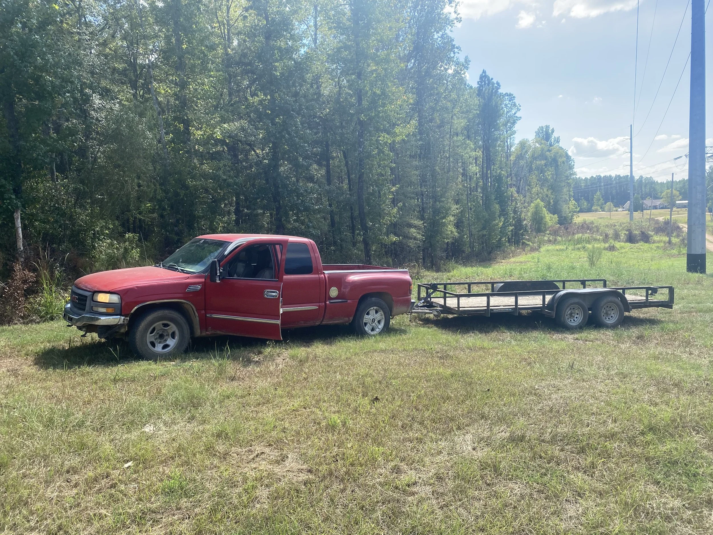 A red pickup truck with an open door, attached to an empty flat trailer, parked on a grassy area near trees and power lines on a sunny day.