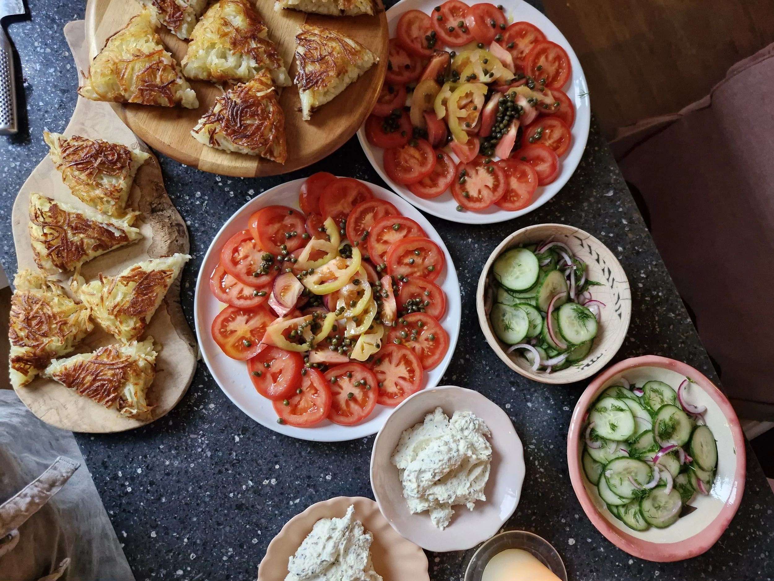 Selection of dishes including tomato salad, cucumber salad, potato wedges with cheese, and cream cheese spread on a black speckled countertop.