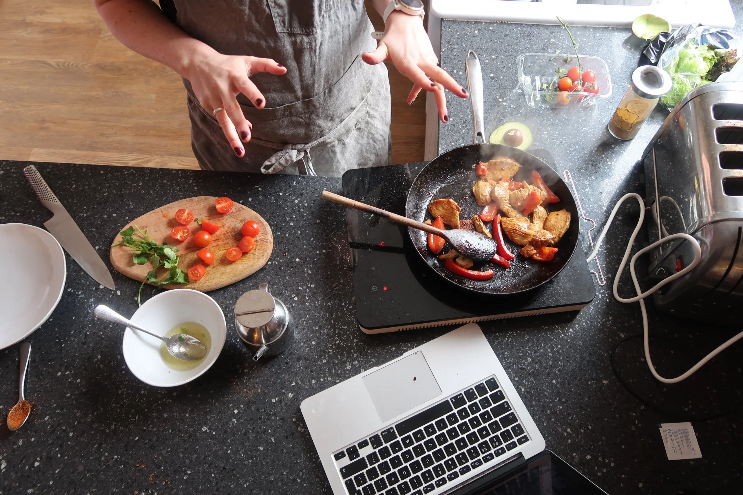 A person cooking chicken and red bell peppers in a black skillet on an electric stove. The kitchen counter has cherry tomatoes, a cutting board with halved cherry tomatoes and fresh herbs, a bowl with olive oil, a small container of spices, and other kitchen utensils.