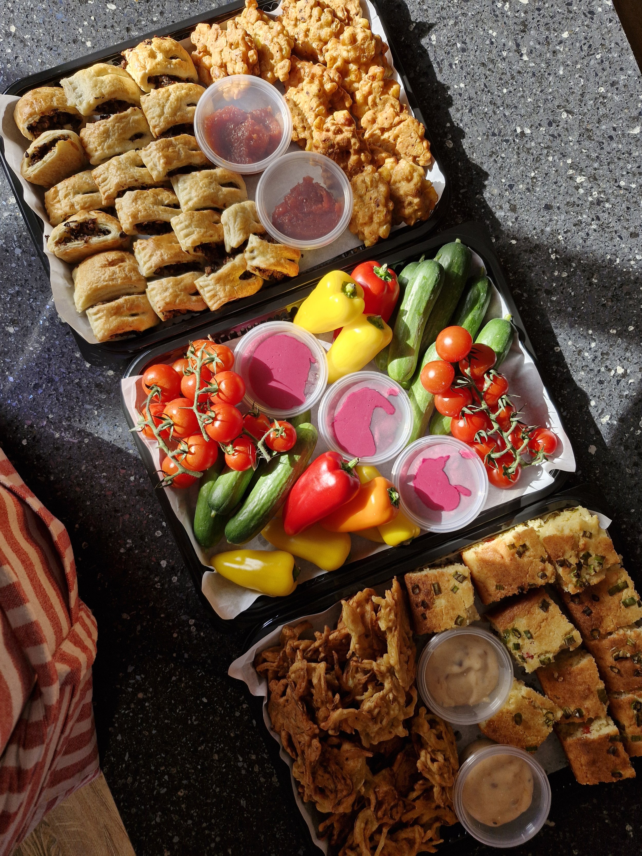 Two catering trays filled with assorted fresh vegetables, dips, and various fried and baked appetizers on a black granite countertop.
