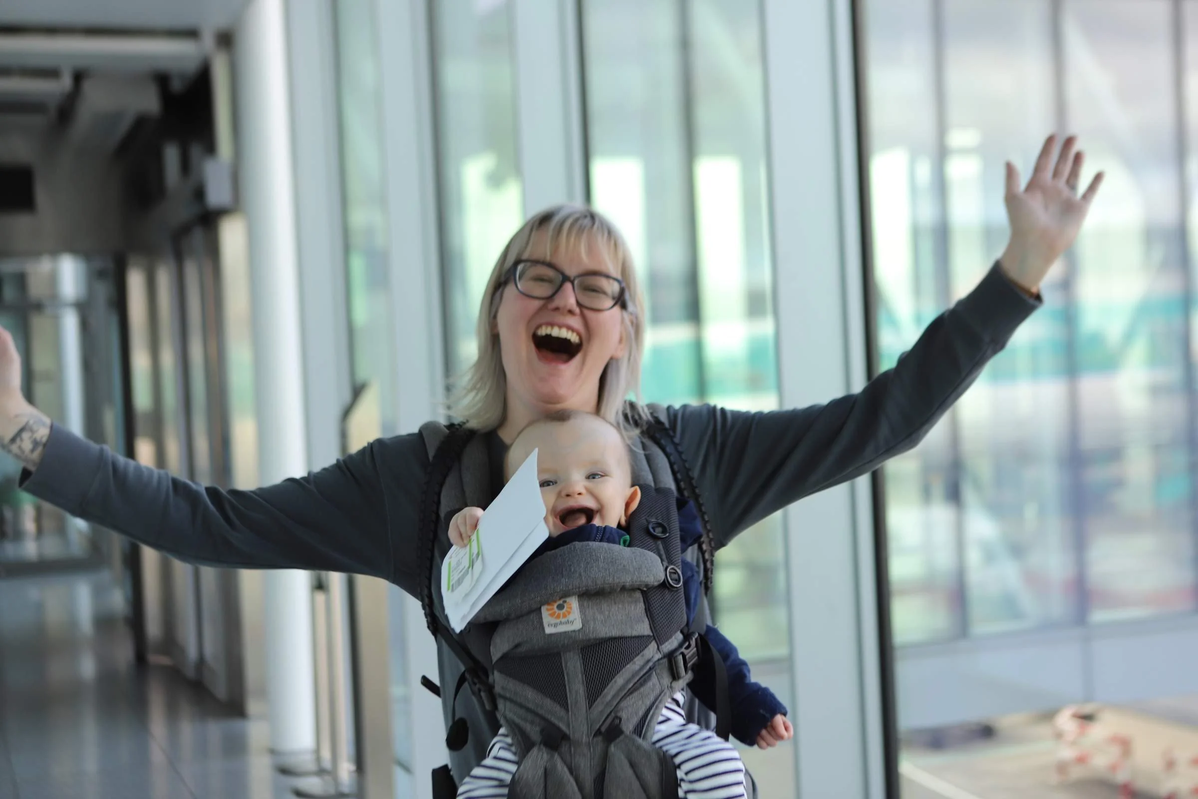 A woman with glasses, smiling and raising her arms, is carrying a baby in a baby carrier. The baby is also smiling and holding a piece of paper, inside an indoor space with glass walls.