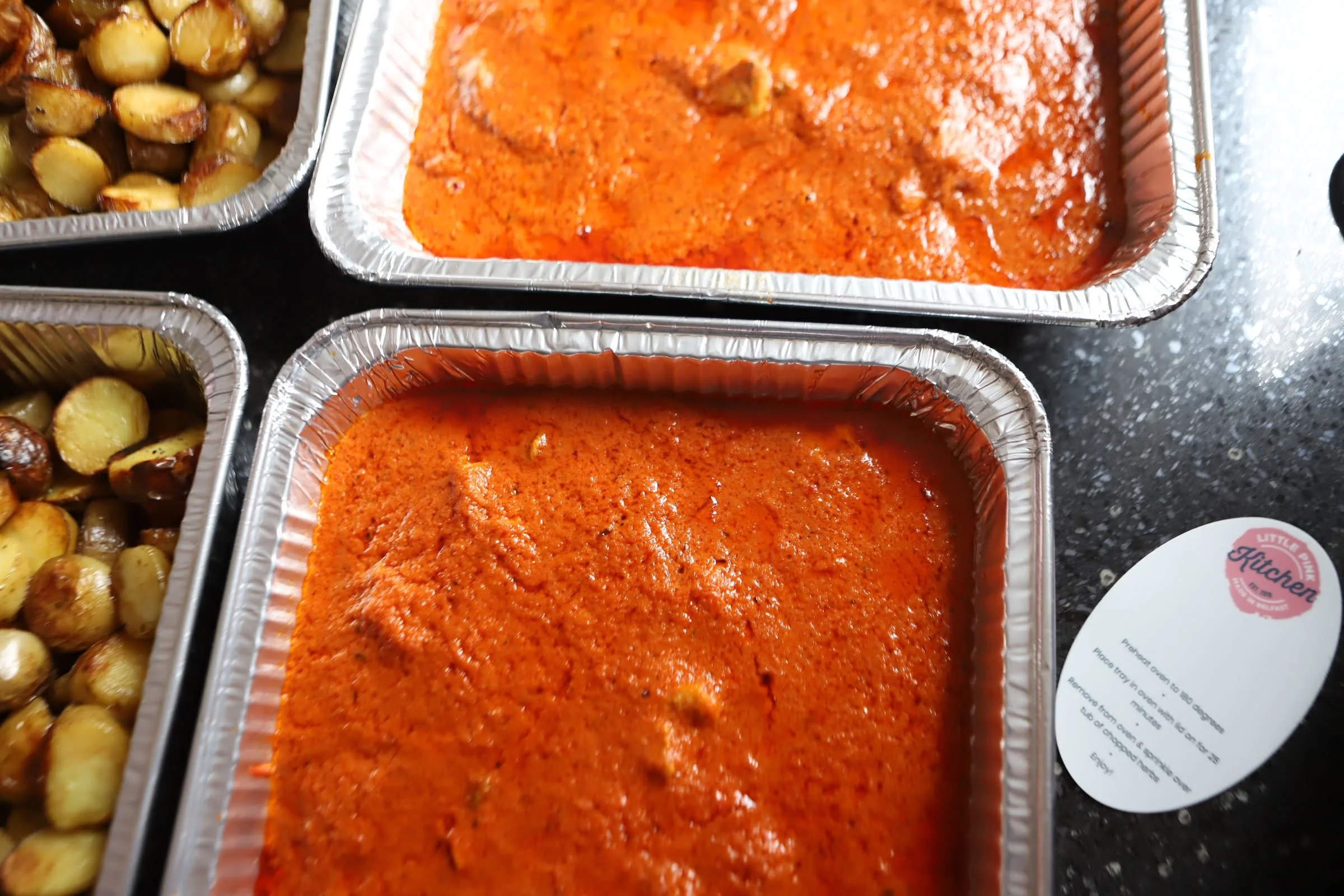 Two aluminum trays of baked chicken in red sauce and two trays of roasted potatoes on a black countertop.