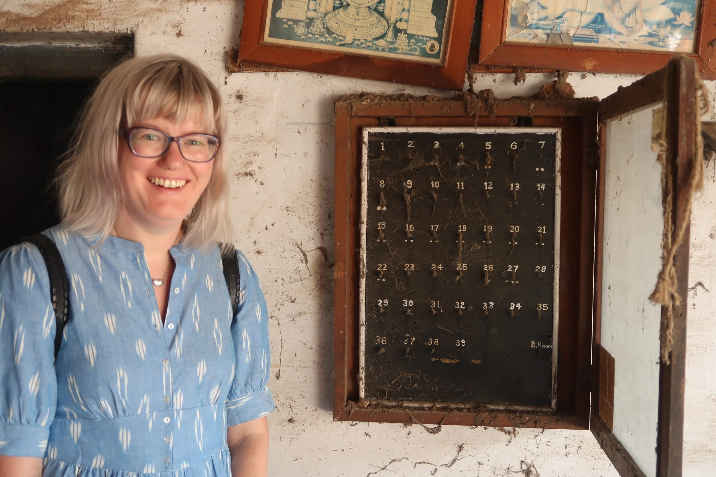 A woman with glasses and blonde hair standing next to an old, dusty, and cobweb-covered blackboard with numbered slots, inside a rustic room with peeling white walls.