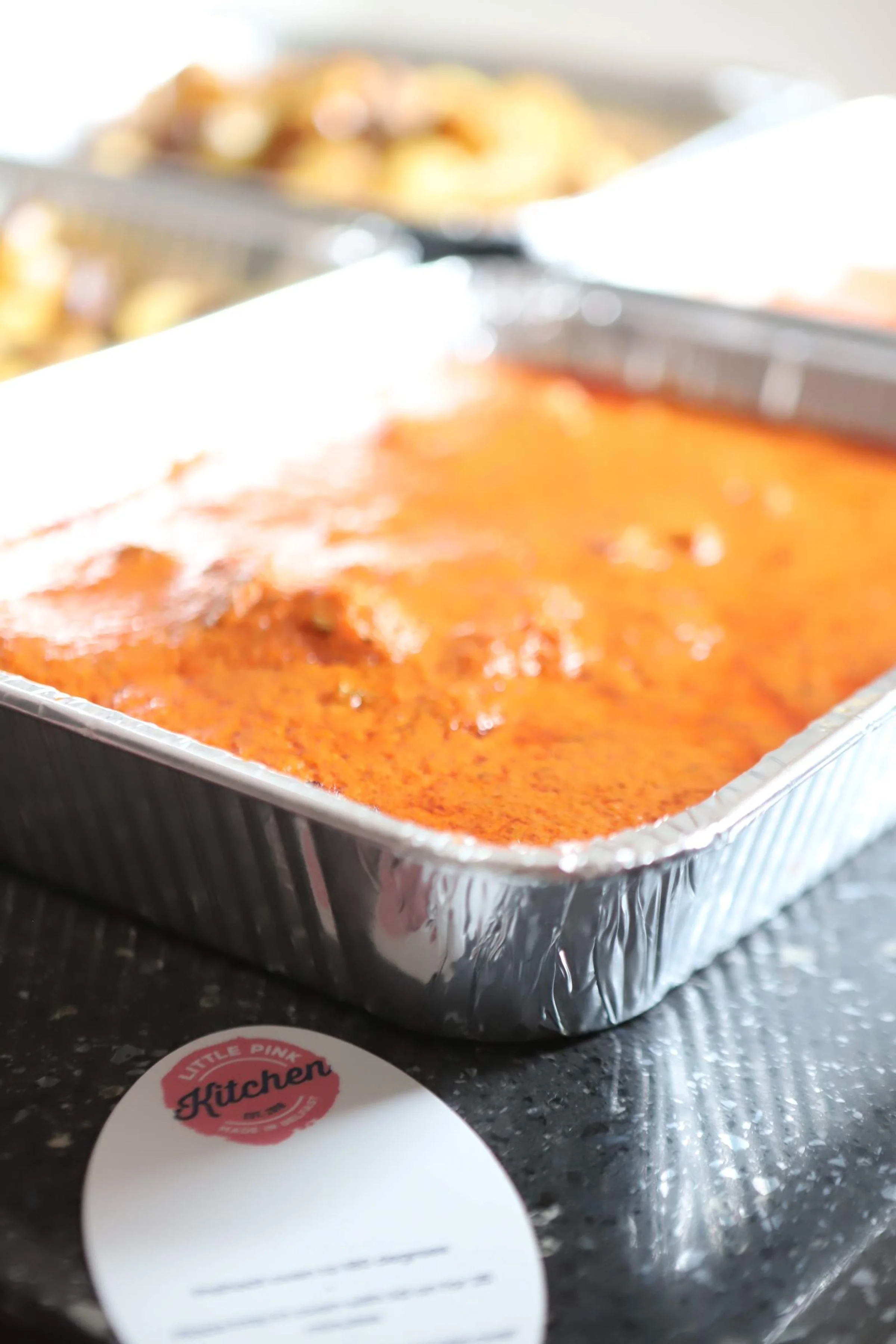Tray of baked lasagna with red sauce, placed on a dark countertop, with a sticker in the foreground that says "Little Pink Kitchen." In the background, out of focus, are other food items or baked goods.
