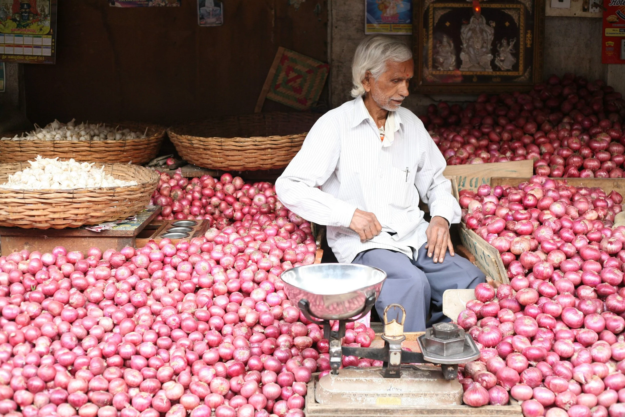 An elderly man sitting amidst a large heap of red onions at a market stall, with wicker baskets and a traditional weighing scale nearby.