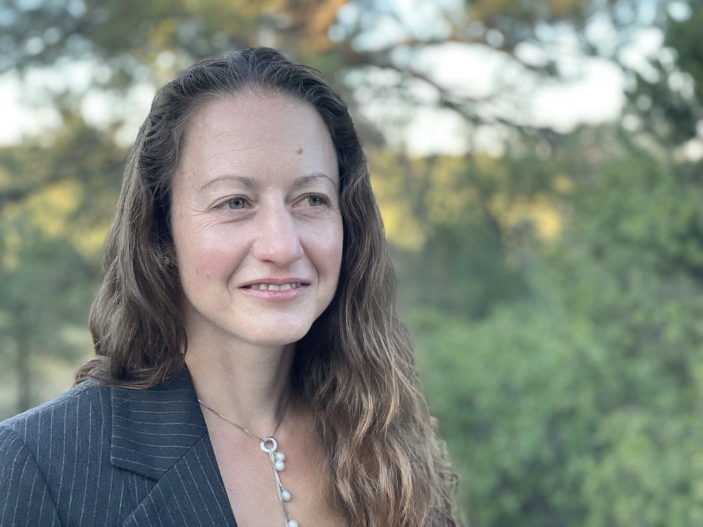 A woman with long wavy brown hair, light skin, wearing a dark pinstripe blazer and a silver necklace, outdoors in front of a blurred background of trees and sky.
