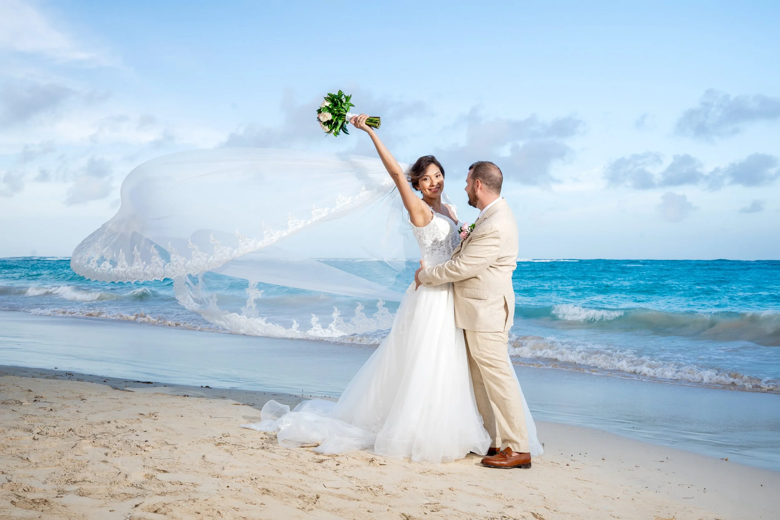 Pareja de recién casados en la playa, ella levantando un ramo de flores y sonriendo, él sosteniéndola por la cintura, fondo de mar y cielo con algunas nubes.