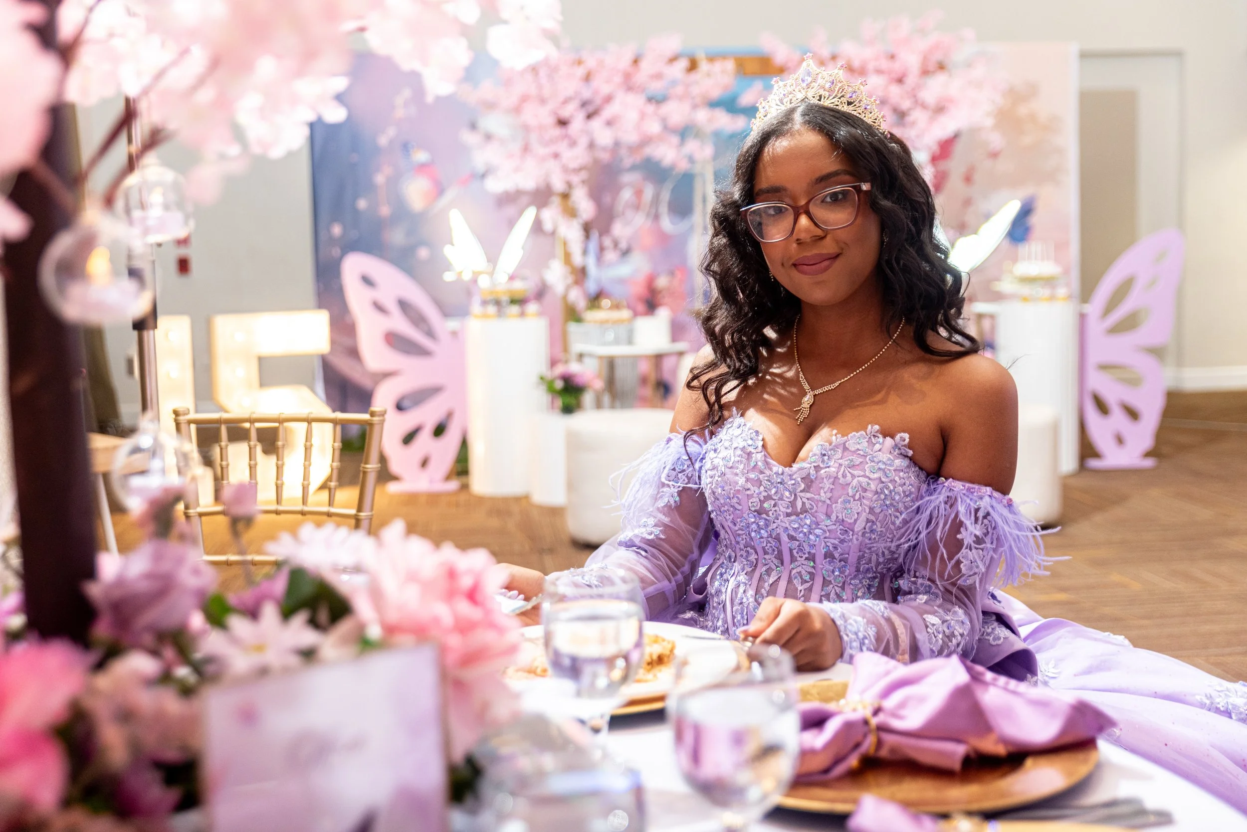 Una joven con vestido lila, corona y lentes, en una fiesta decorada con temática de mariposas y flores rosadas.