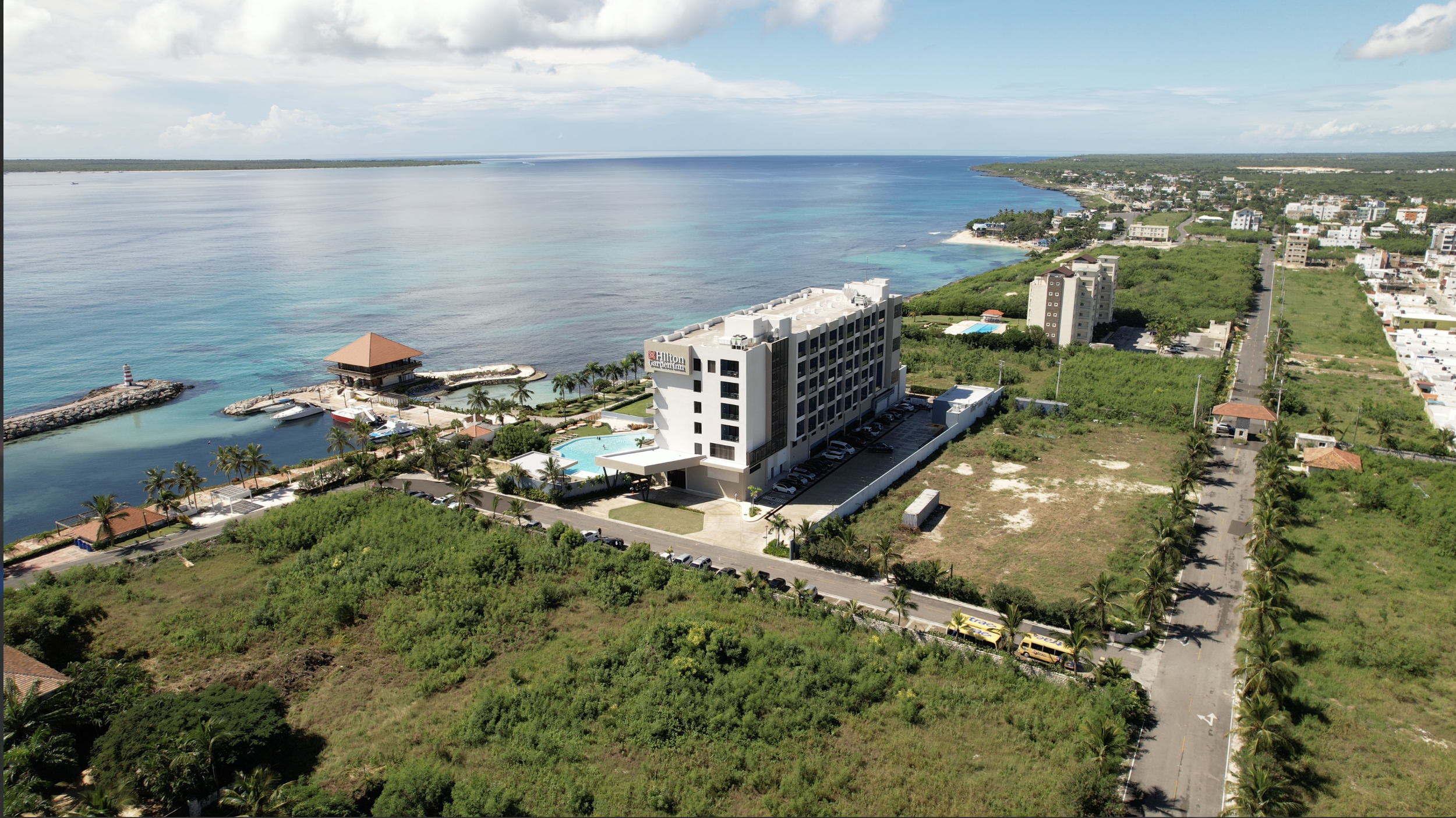 Vista aérea de un hotel Hilton en una zona costera con playa y mar turquesa, jardines con palmeras, área de piscina, zona de muelle con embarcaderos y una ciudad pequeña alrededor.
