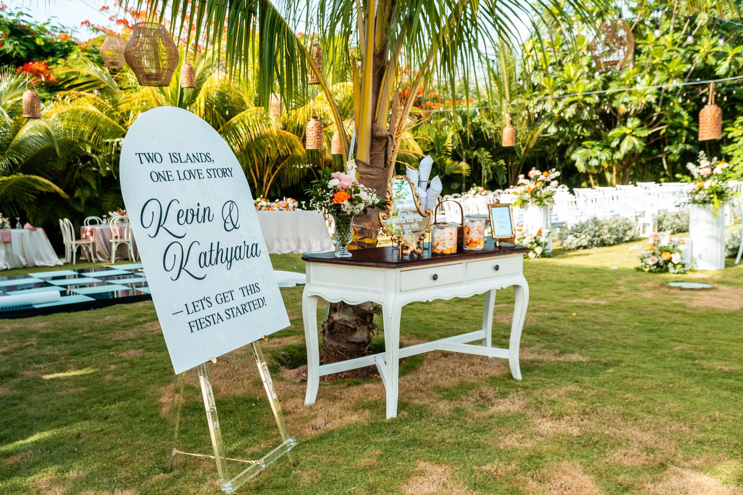 Decoración de boda en jardín con una mesa blanca decorada con flores, un cartel de bienvenida con los nombres Kevin Q y Kathya y una señal que dice "Let's get this fiesta started!"
