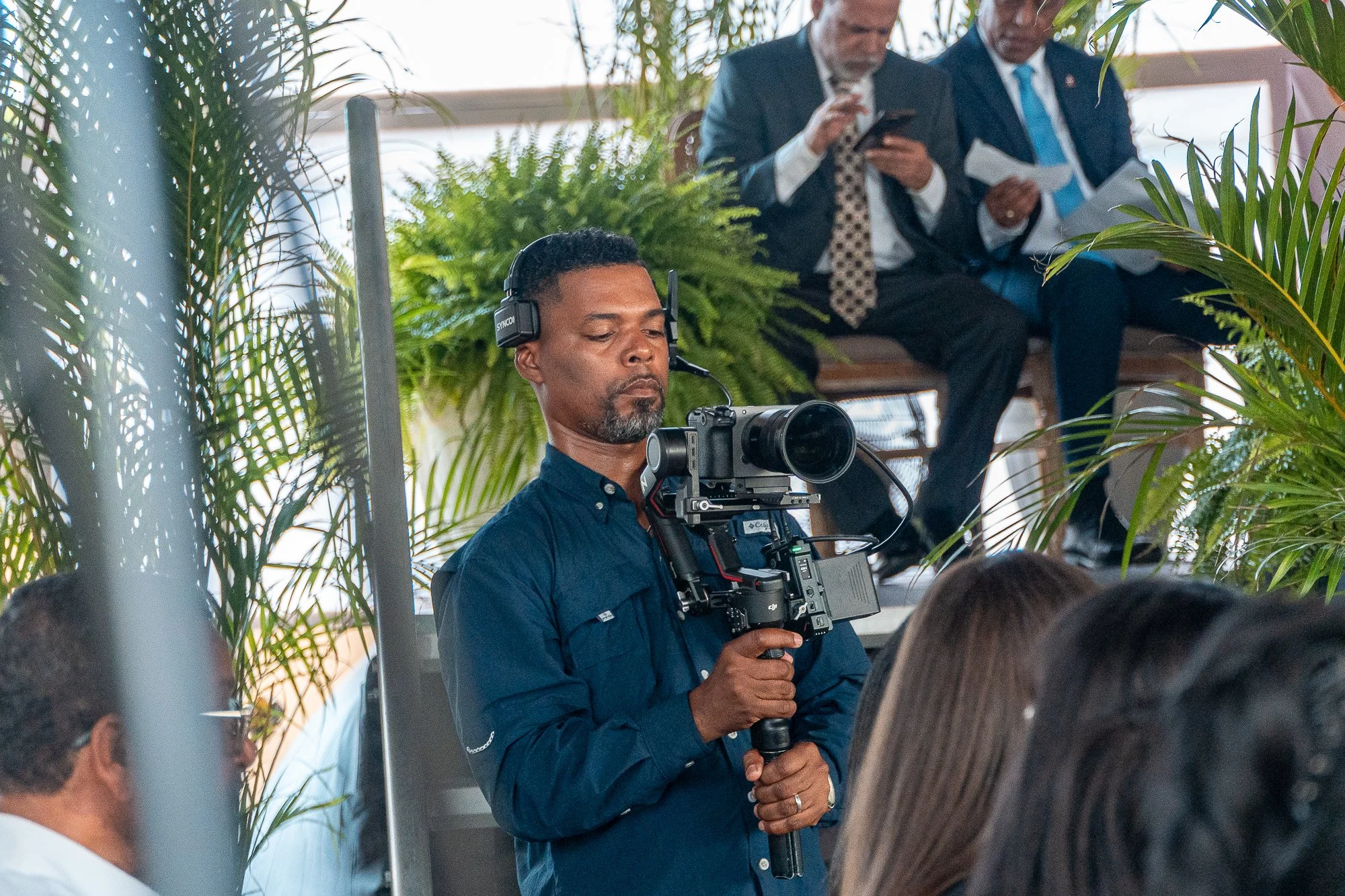 Un camarógrafo grabando en un evento con varias personas sentadas y un fondo con plantas verdes.