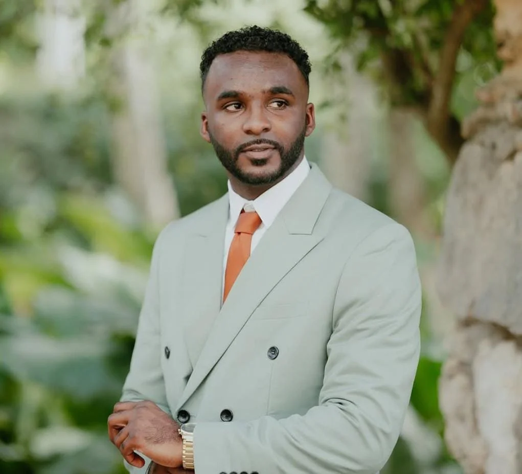 Hombre de negocios con traje gris, camisa blanca y corbata marrón, en un entorno natural con árboles y piedra.