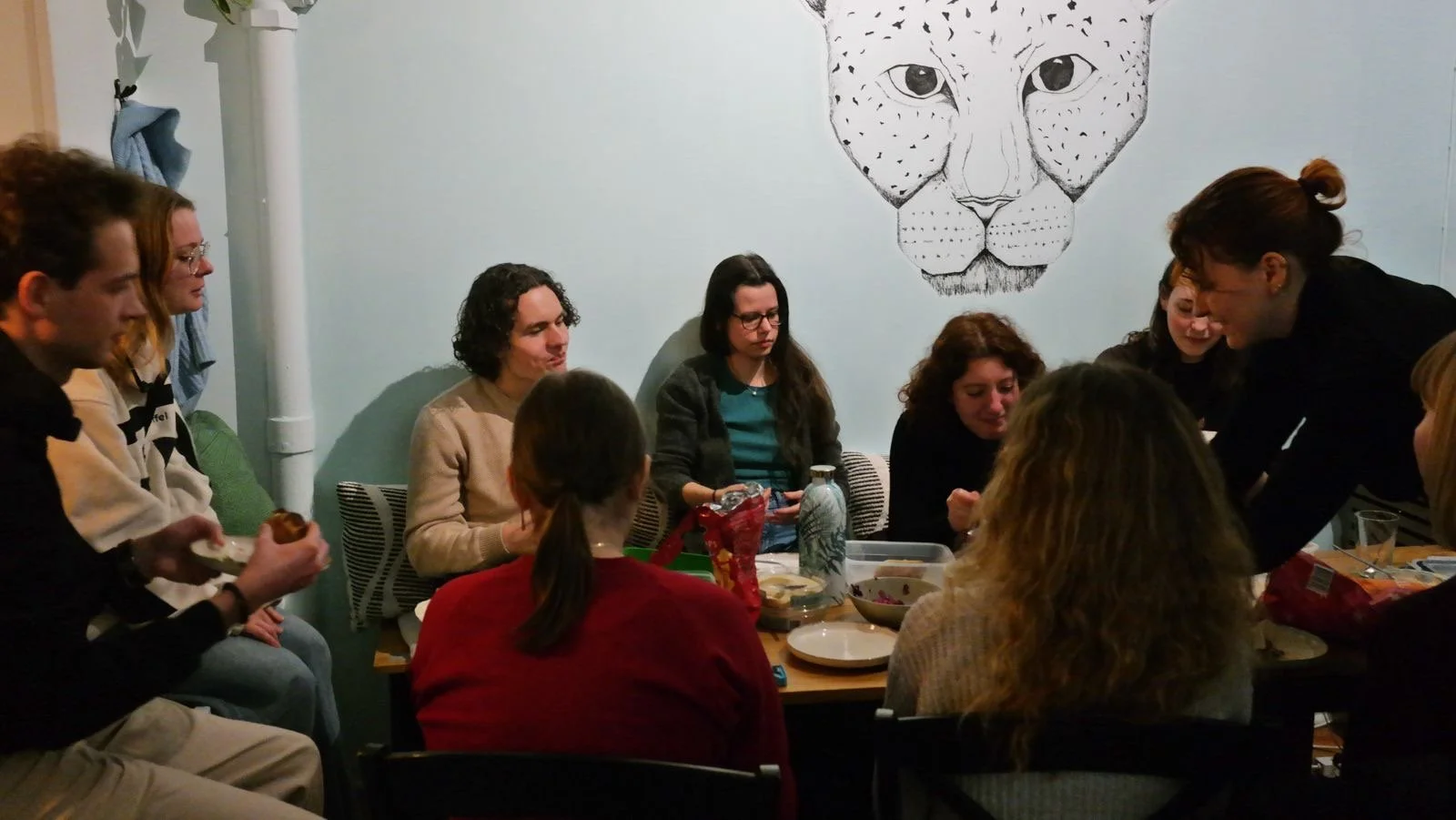 Group of people gathered around a table, some standing and some sitting, with food and drinks, in a room with a large drawing of a leopard's face on the wall.