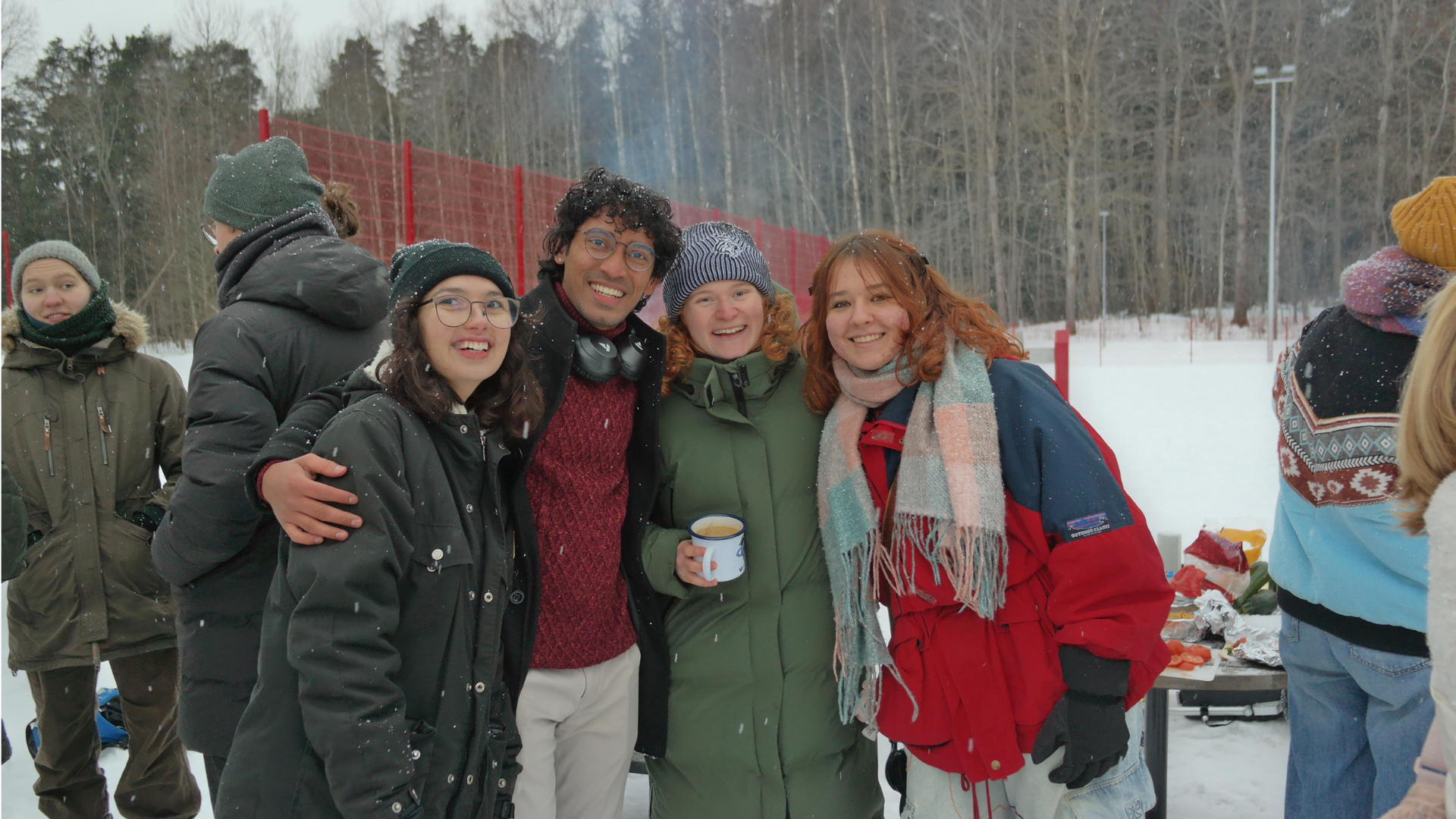 Group of five friends standing outdoors in a snowy area, smiling and bundled up in winter clothing, with a table of food and snacks behind them.