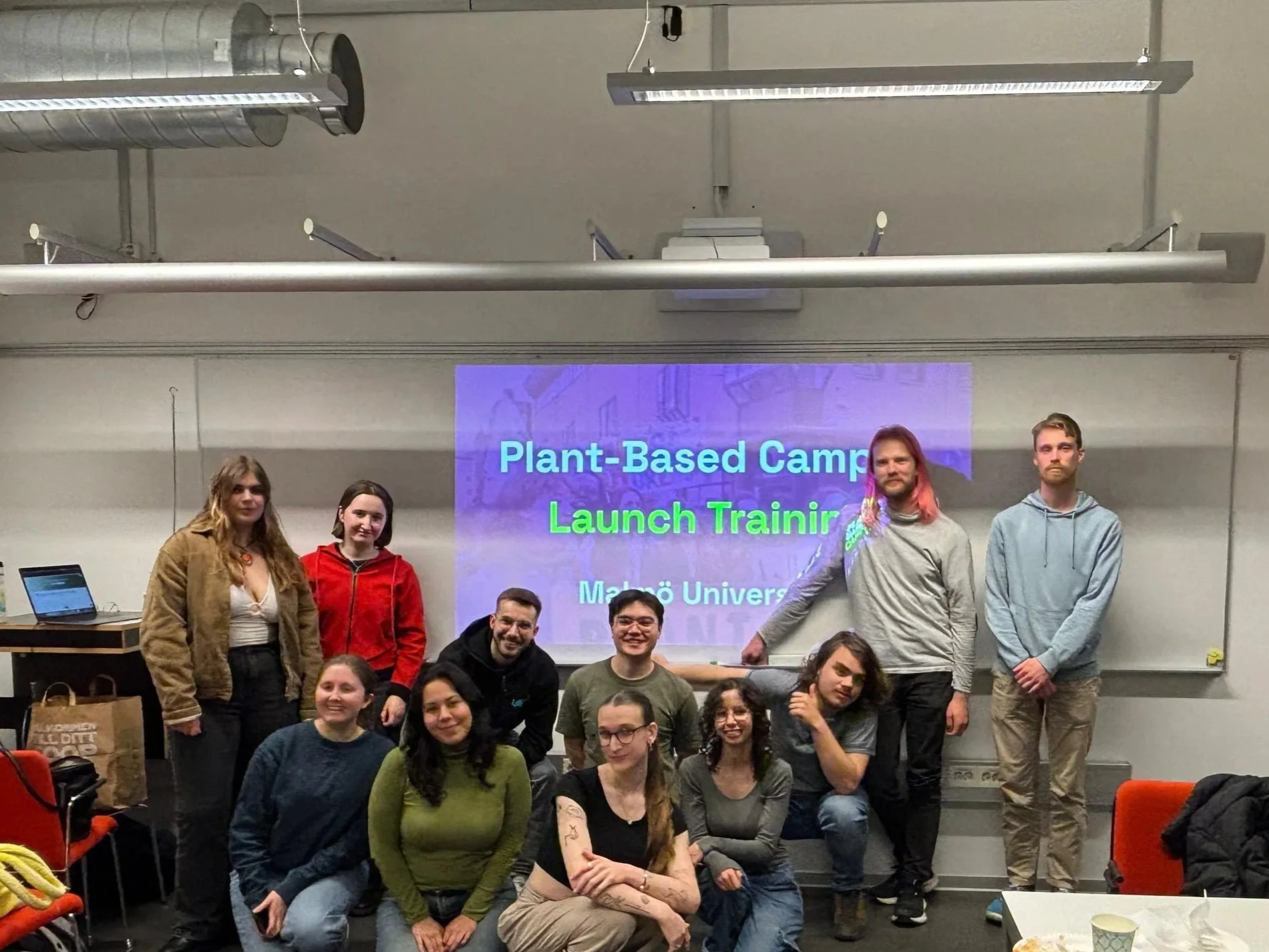 Group of people gathered in front of a presentation screen that reads 'Plant-Based Camp Launch Trainer' at Malmö University, posing for a photo in a classroom or conference room.