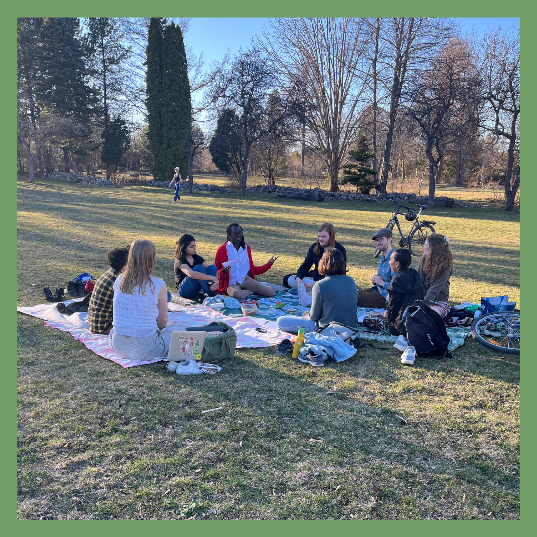 Group of people sitting on a blanket having a discussion in a park with trees and a bicycle in the background.