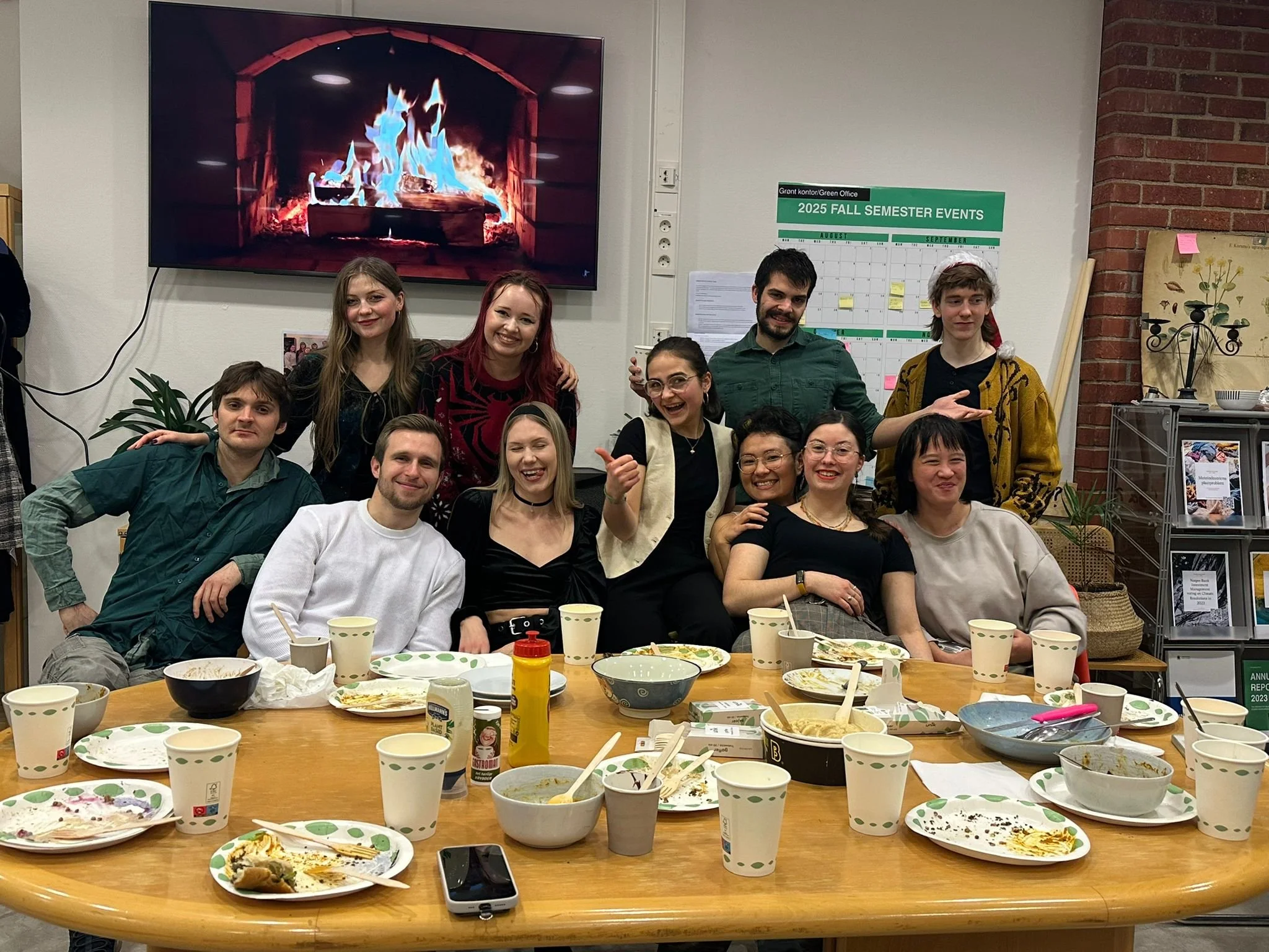 Group of people gathered around a table with leftover food, paper cups, and plates, celebrating at an indoor event with a fireplace on a large screen in the background.