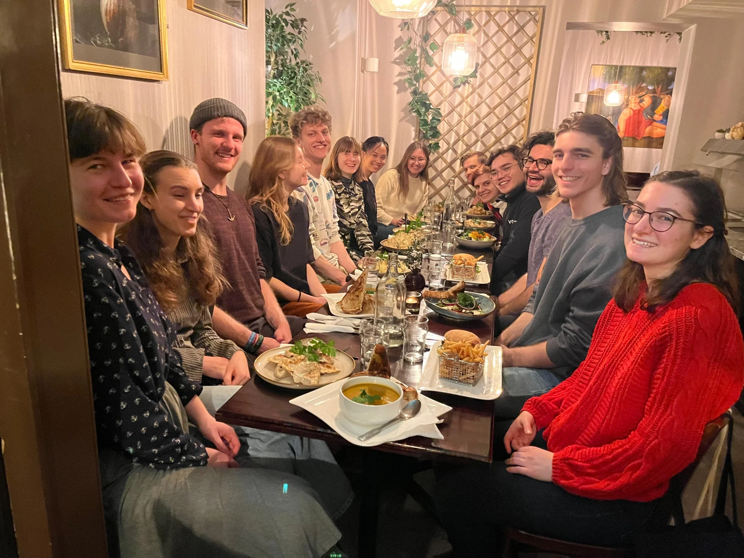 A group of people sitting at a long dining table in a restaurant, smiling and enjoying a meal together. The table is set with plates, glasses, pitchers of water, and various dishes. The restaurant has warm lighting with framed artwork on the walls and decorative greenery.