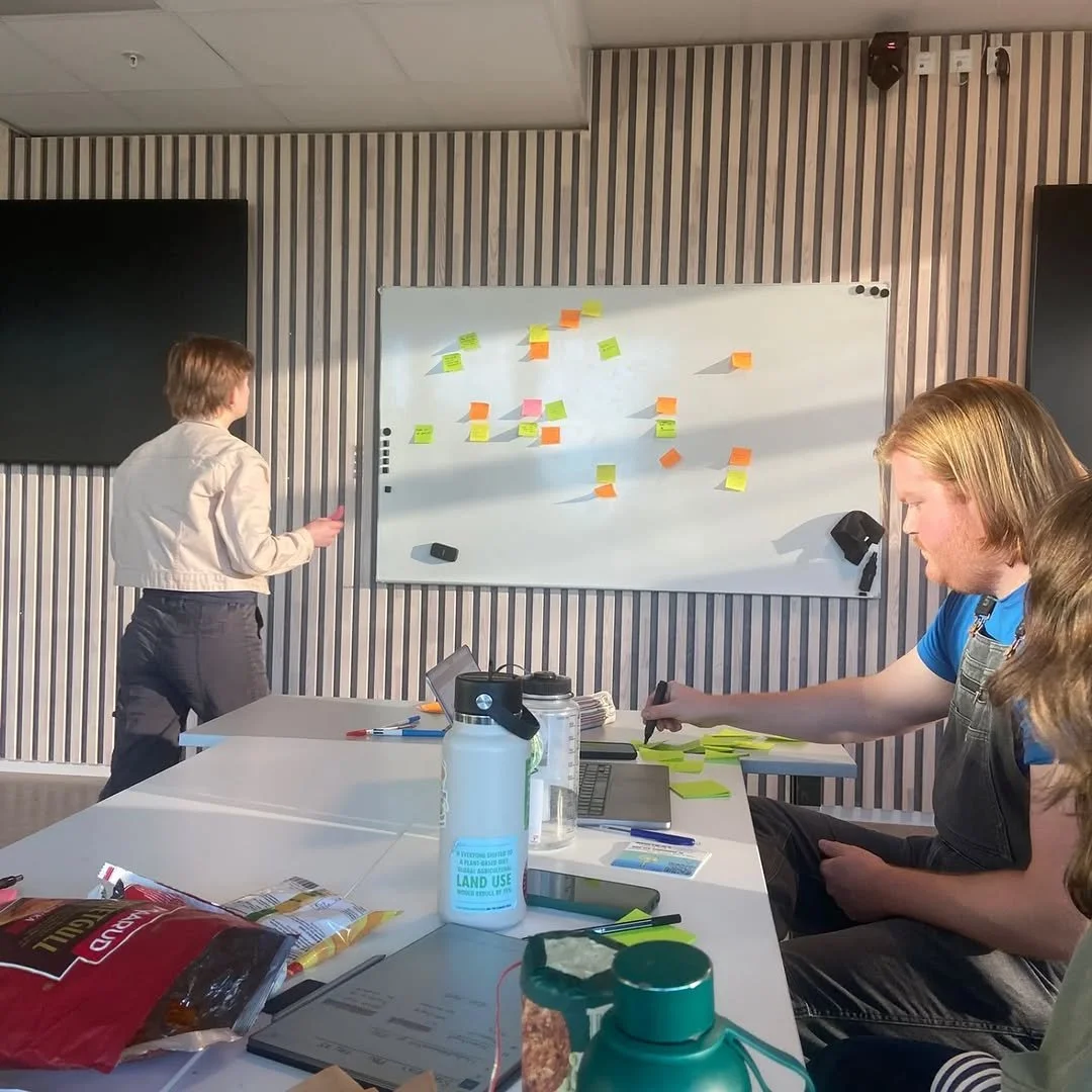 A group of people in a meeting room with a whiteboard covered in colorful sticky notes, some people are seated and one person is writing on the sticky notes. There are snacks and drinks on the table.