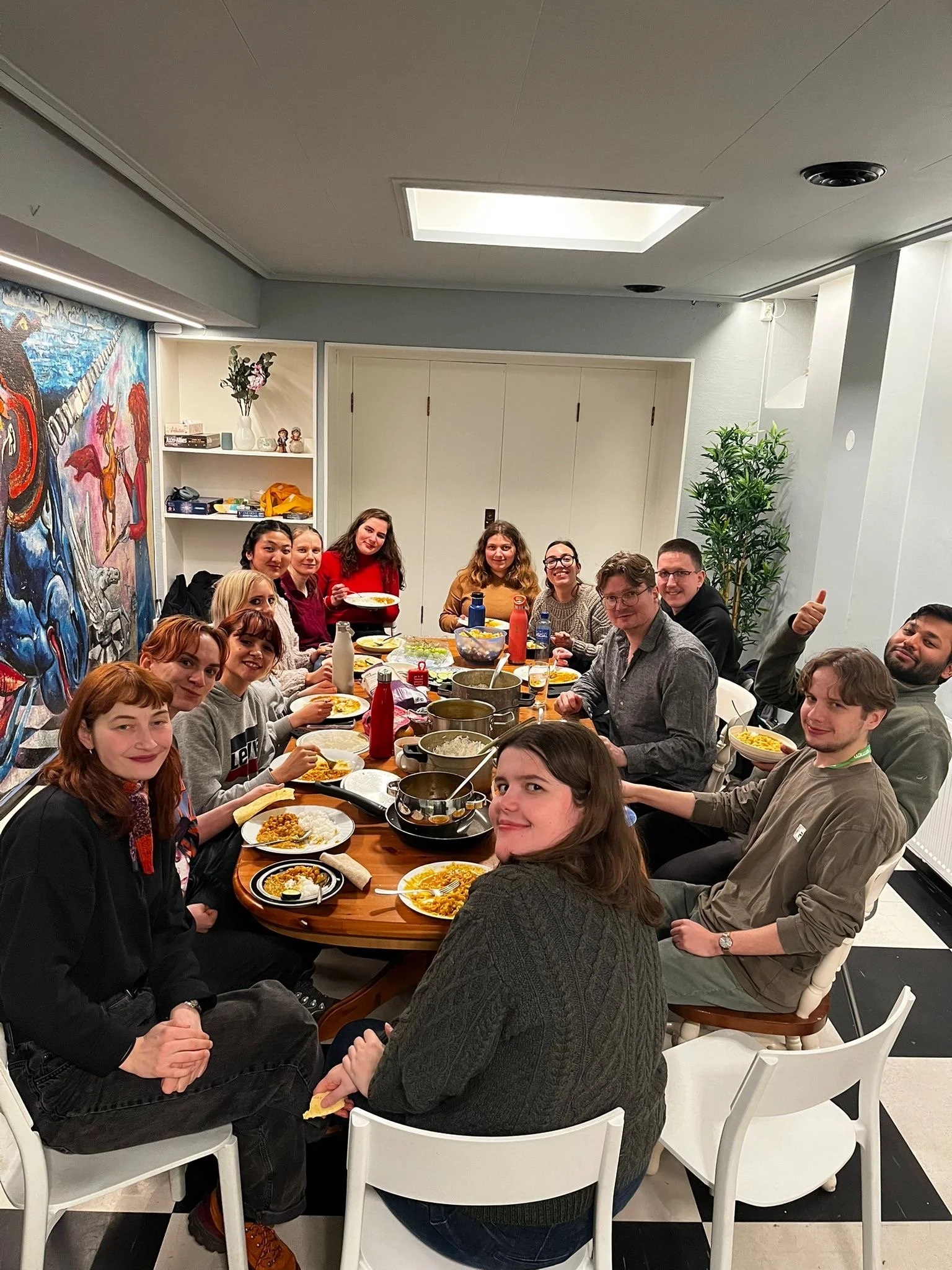 Group of people sitting around a dining table enjoying a meal in a well-lit room with colorful artwork on the wall and a tall green plant in the background.