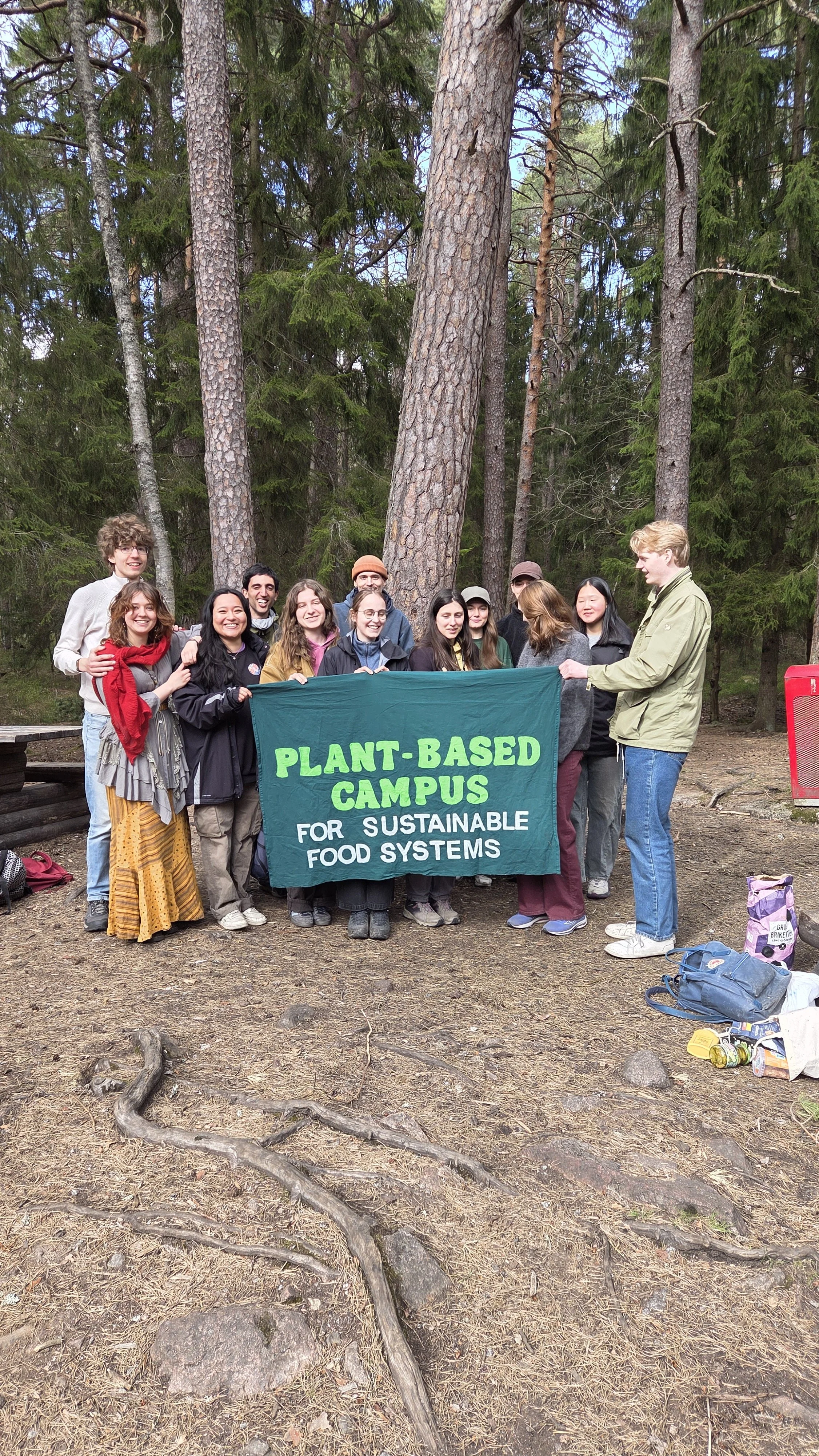 A group of people outdoors in a forest, holding a green and white banner that reads "Plant-Based Campus for Sustainable Food Systems." They are smiling and standing among tall trees and natural ground cover.