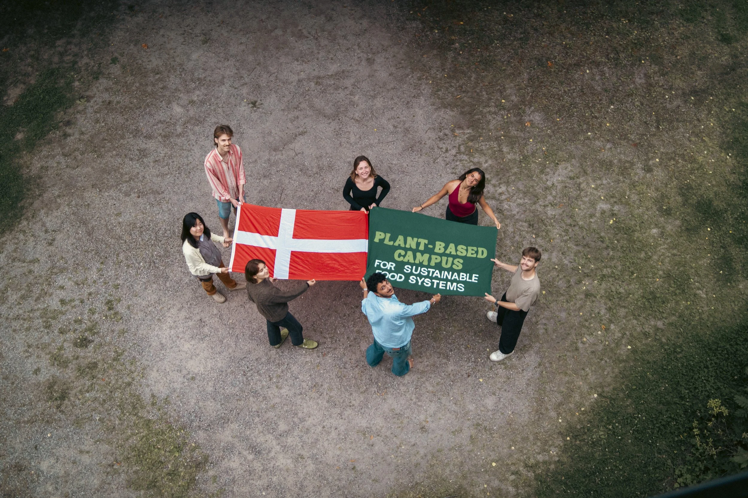Six people standing in a circle outdoors, holding a Danish flag and a green banner that reads 'Plant-Based Campus for Sustainable Food Systems'.