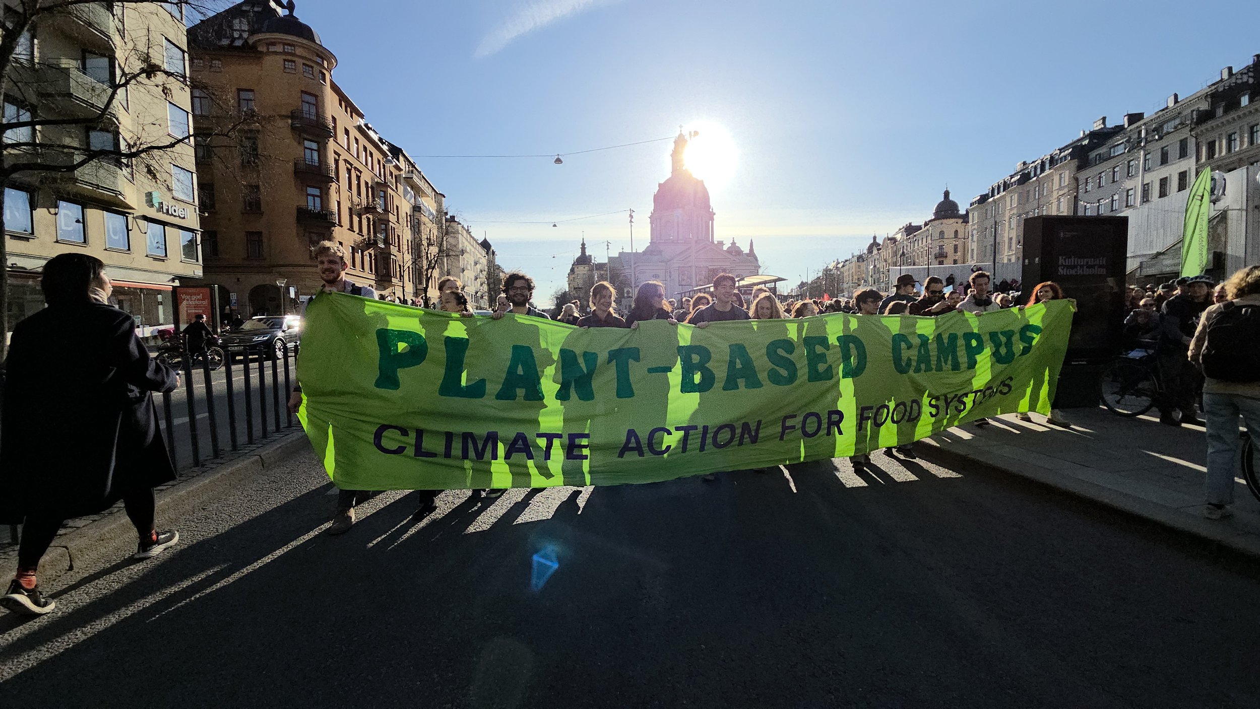 People marching in a city street carrying a large green banner that reads 'Plant-Based Campus Climate Action for Food Systems' with historic buildings and a church tower in the background, and the sun shining behind the tower.