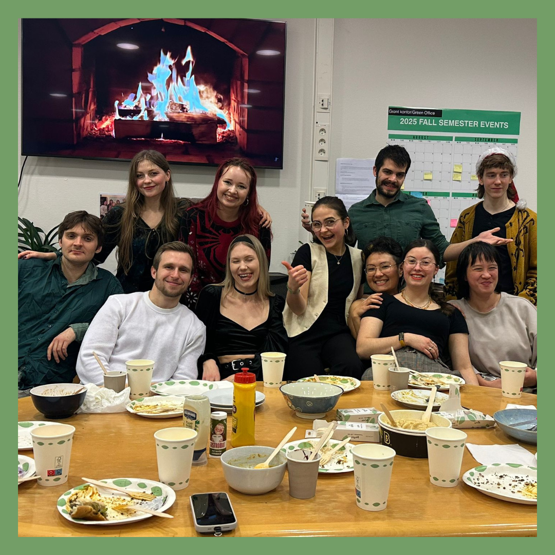 Group of ten people gathered around a table with leftover food and drinks, smiling and posing for a photo in an indoor setting with a large fireplace image on a TV screen behind them and a calendar on the wall.
