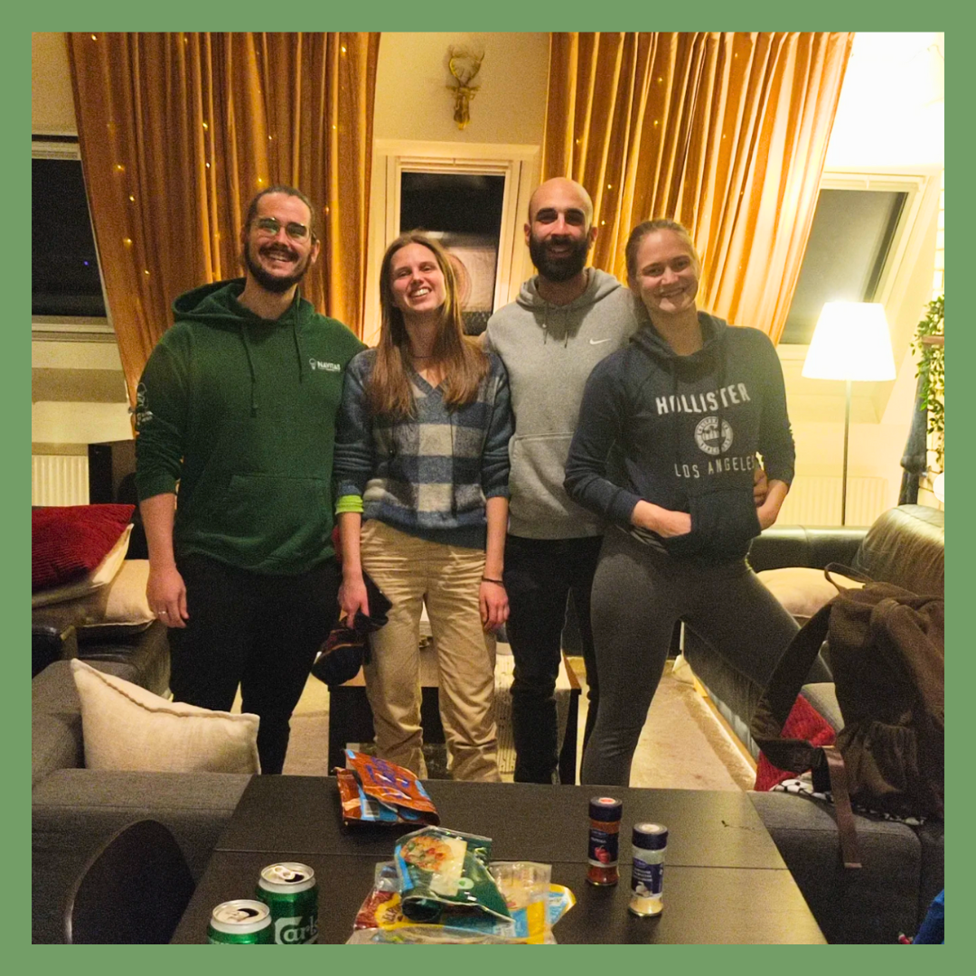 Four people standing in a living room, smiling at the camera. They are behind a table with snacks and drinks, with a window and patterned curtains in the background. The room has warm lighting and cozy furniture.