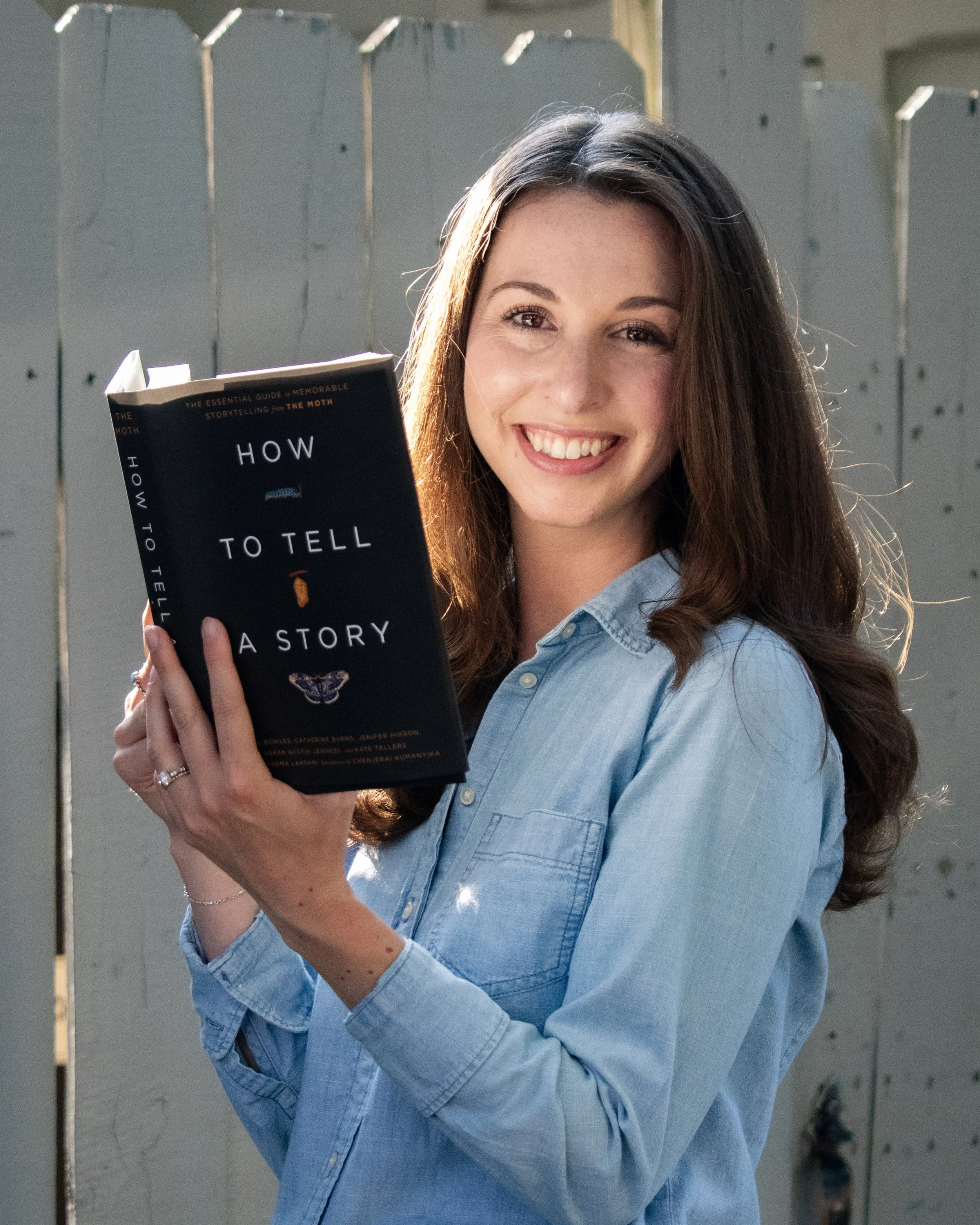 A smiling young woman with long brown hair holding a book titled 'How to Tell a Story' near a gray wooden fence during daylight.