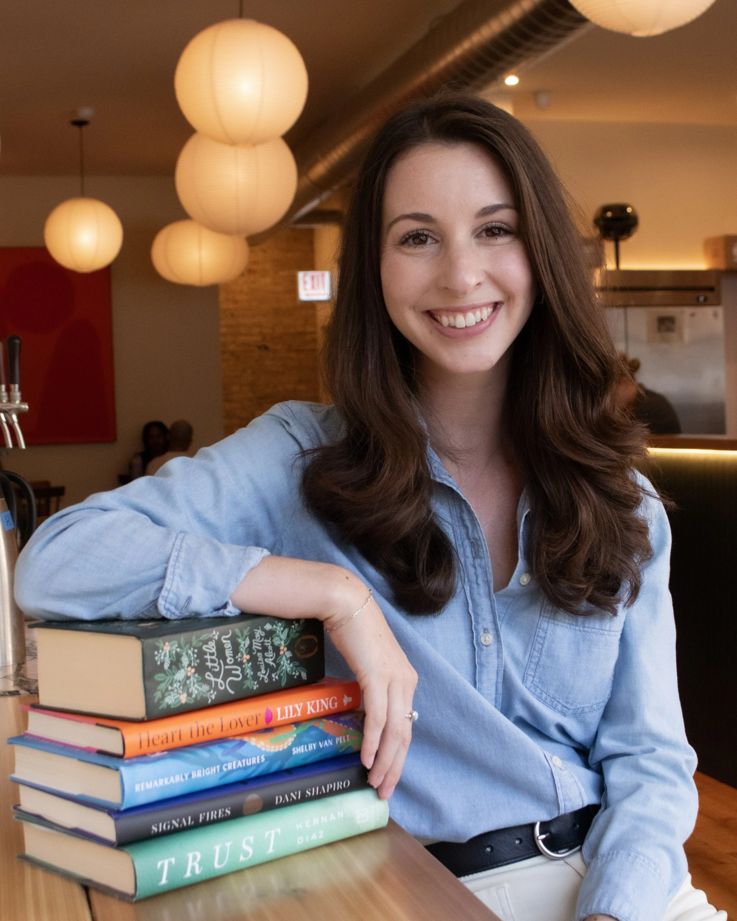 A smiling woman with long brown hair sitting at a table in a restaurant, with a stack of colorful books on the table, including titles like "Little Women" and "Heart the Lover".