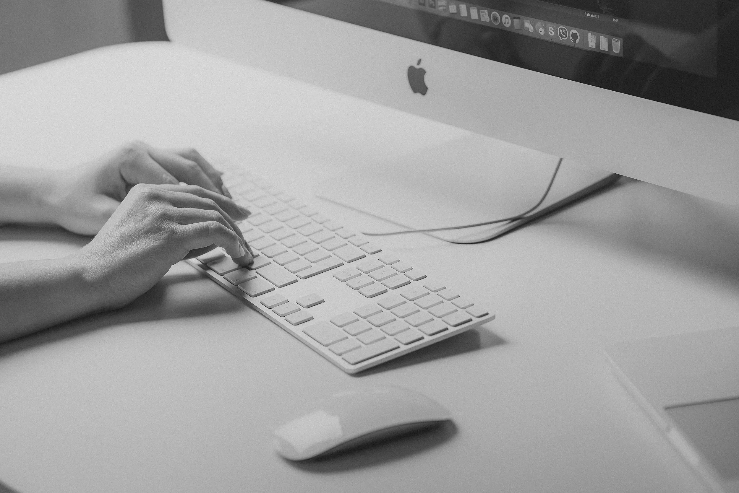 Black and white photo of a person typing on an Apple wireless keyboard with an iMac computer in the background on a white desk.
