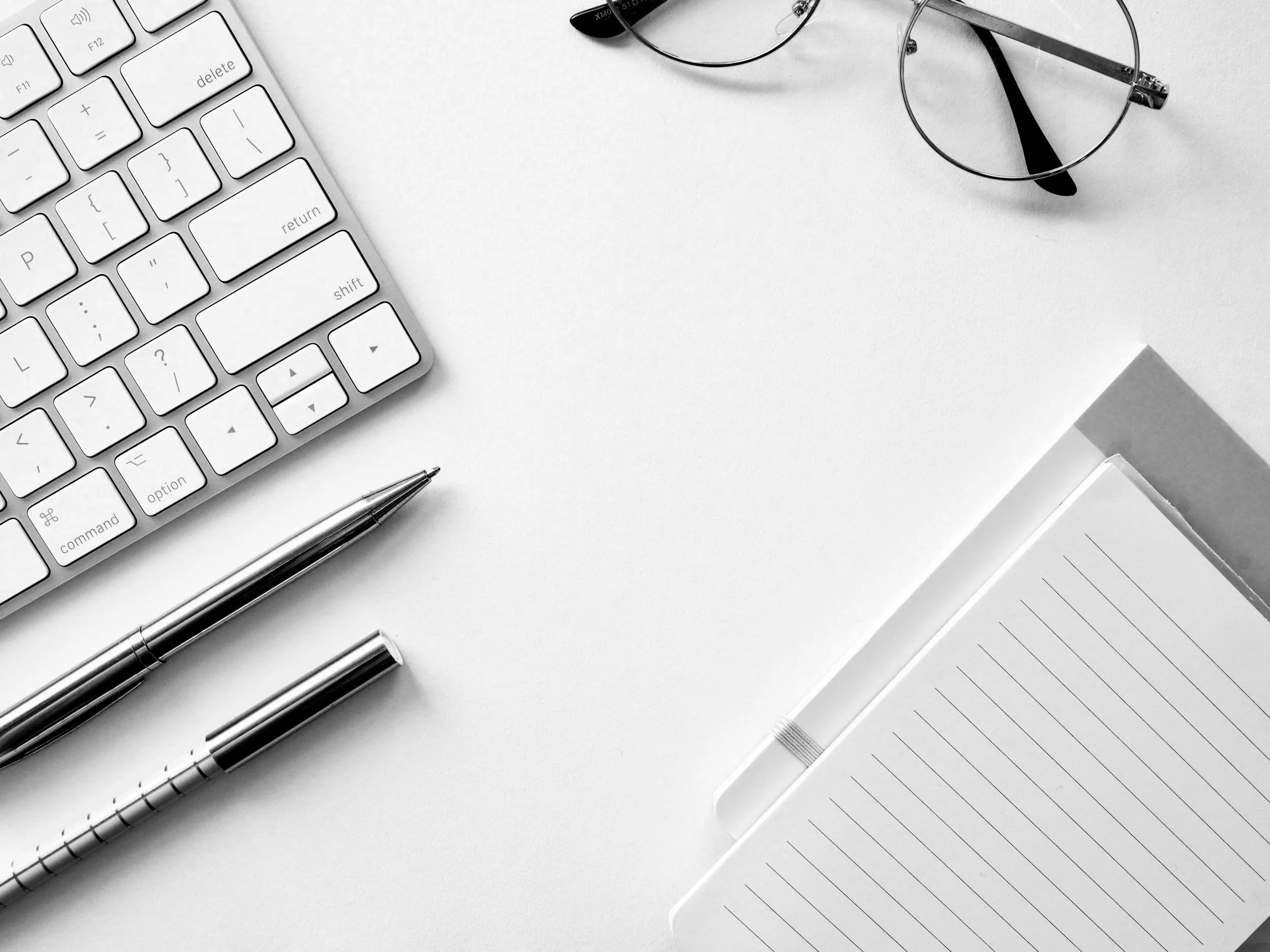 Desk with white keyboard, silver pens, eyeglasses, notepad, and a box on white surface.