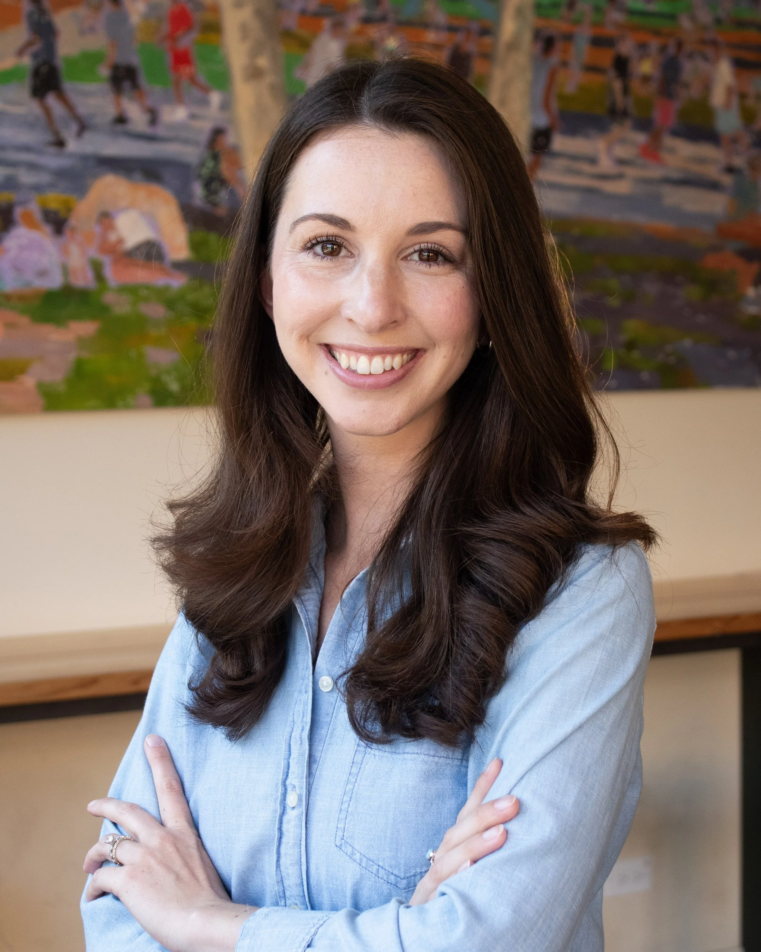 A woman with long brown hair wearing a light blue button-up shirt, standing with arms crossed in front of a colorful painting.
