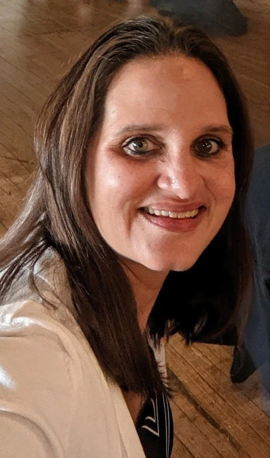 A woman with brown hair and light skin smiling at the camera, wearing a white top, indoors with wooden flooring.