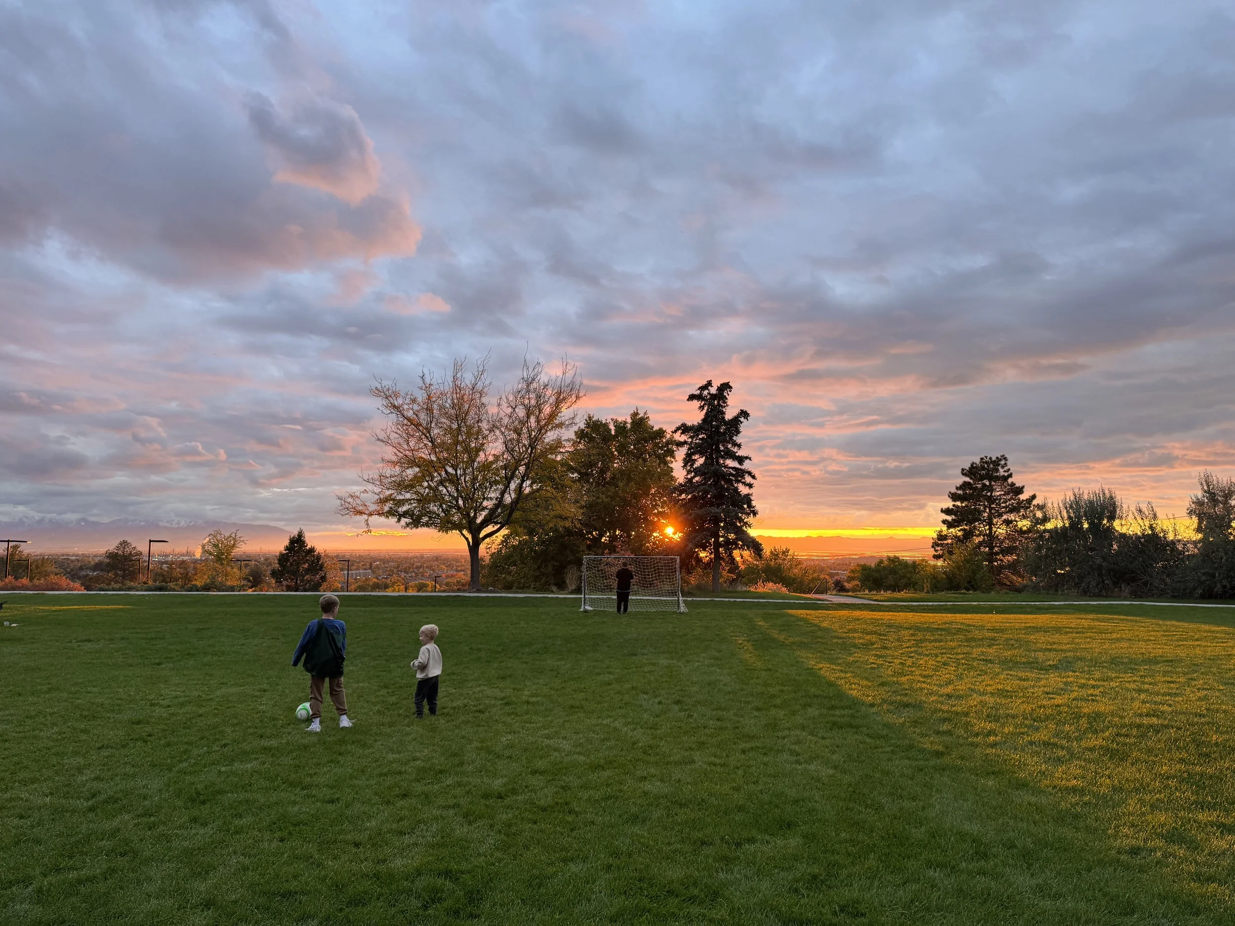 Two children playing soccer on a field at sunset, with trees and a cloudy sky in the background.