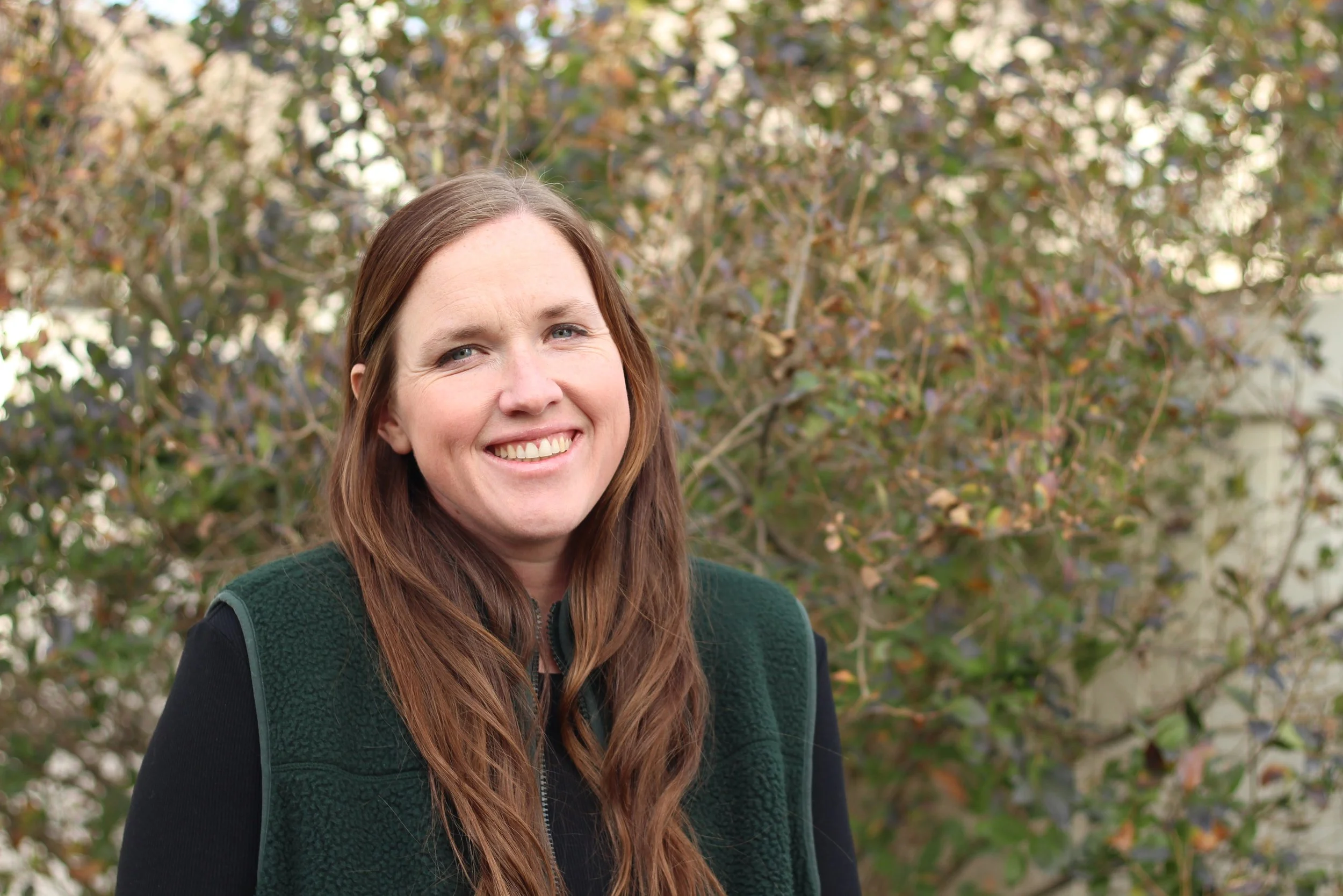 A smiling woman with long brown hair standing outdoors in front of a bush with green and brown leaves.